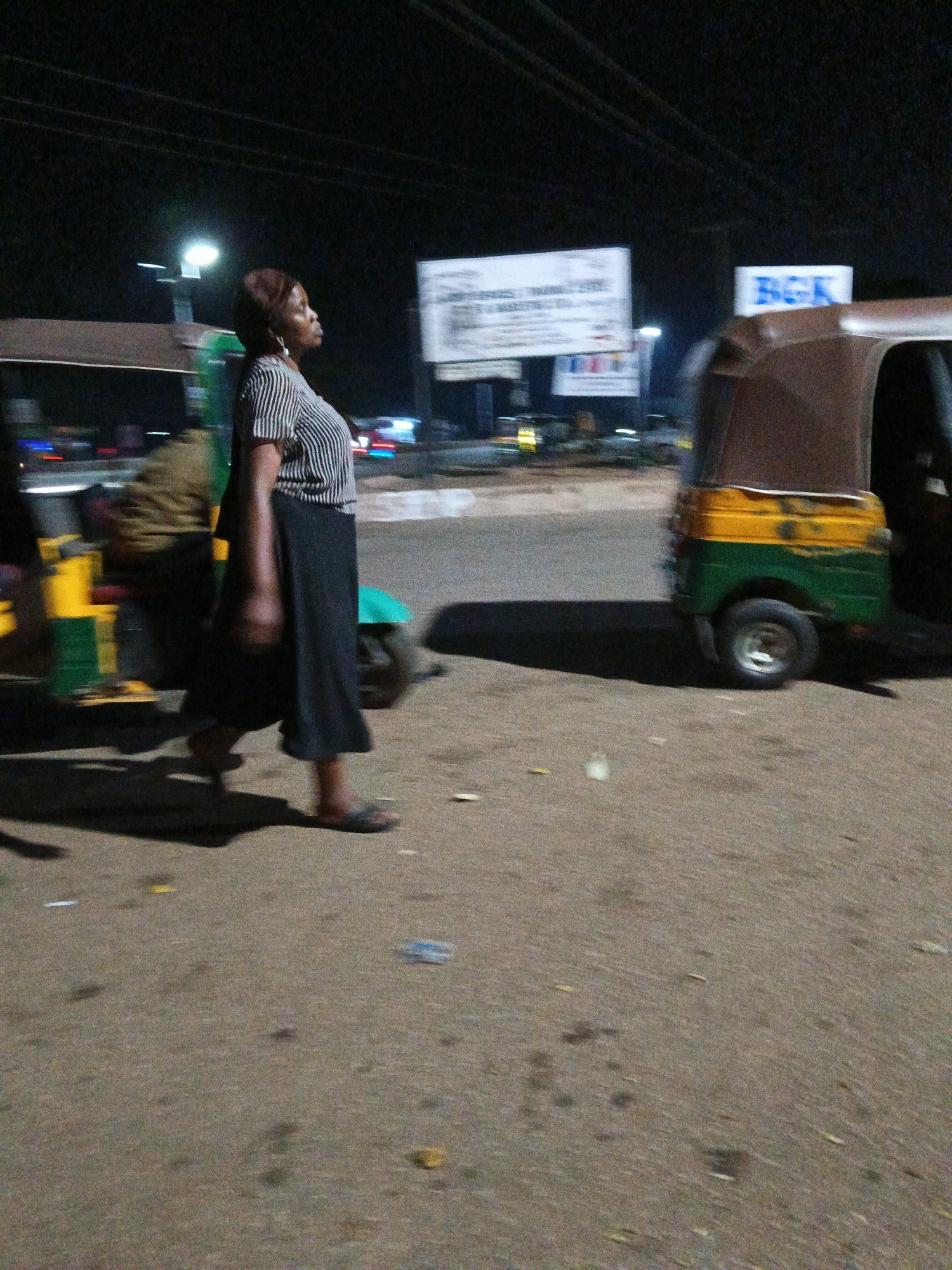An over-the-shoulder shot shows a Black woman in a striped shirt and long black skirt walking to the left in Bukuru, Nigeria. She has a dark headwrap and a large hoop earring in her left ear. She is walking on a paved surface next to a multi-colored tuk-tuk, partially obscured by the woman. The tuk-tuk is green and yellow. In the background, blurred lights and signs indicate it is nighttime. One sign in the background reads "BGK" in large blue letters. Another, larger sign is white with black text. The overall scene is dimly lit, suggesting it is evening or night.