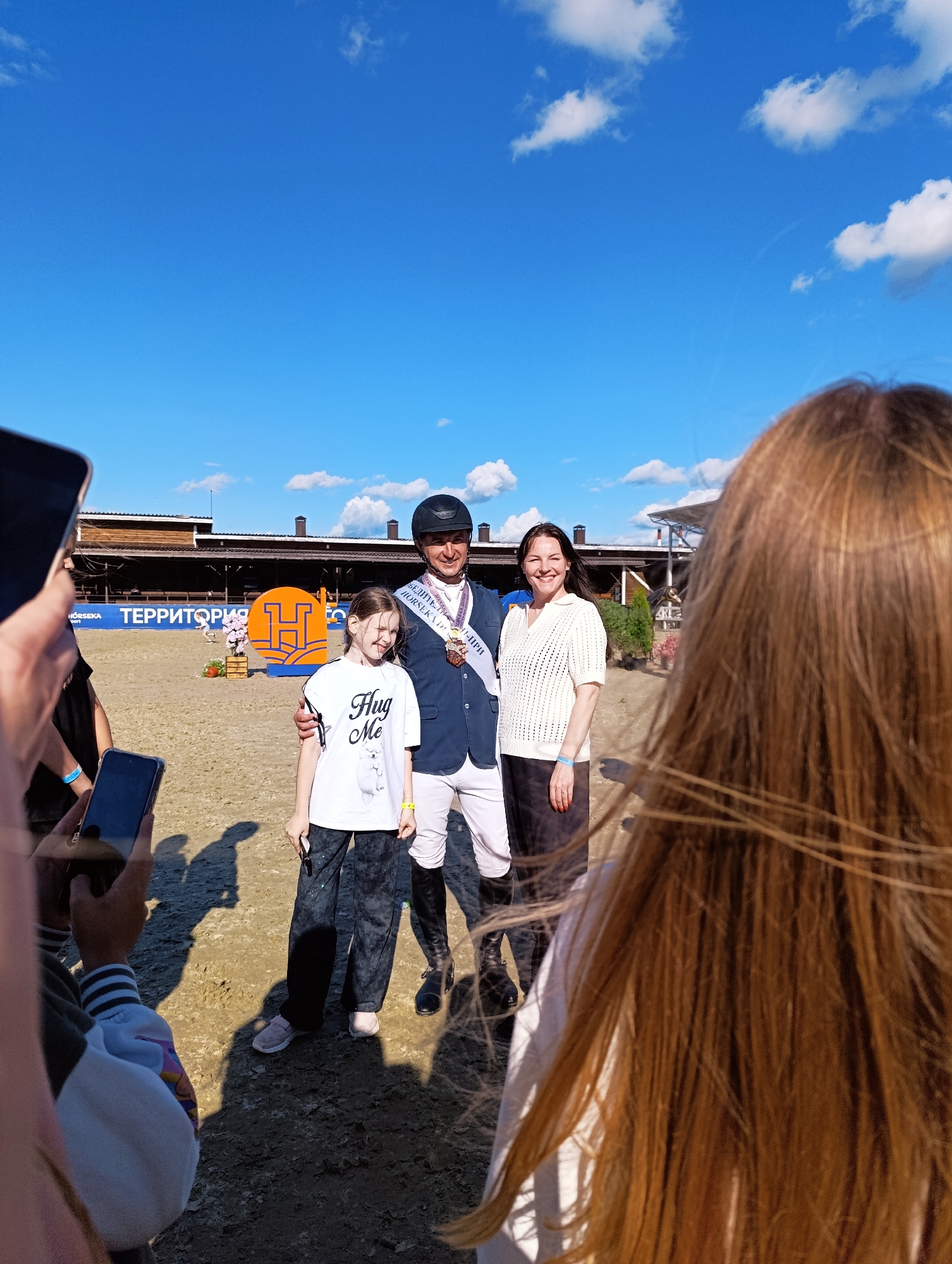 A group of people are posing for a photo on a sandy surface, likely an equestrian arena, given the backdrop of wooden bleachers and a large orange "H" logo, possibly representing a venue or sponsor. The sky is a clear, bright blue with scattered white clouds, suggesting it is daytime and the weather is fair.
In the center of the group is a man dressed in a dark blue equestrian jacket, white riding pants, and a black helmet. He is wearing a sash that appears to be ceremonial and has a medal around his neck, indicating he may have won an award or participated in a competition. He is smiling warmly.
To the man's left is a young girl wearing a white t-shirt with the words "Hug Me" and a graphic of two animals. She is also smiling and looking towards the camera. To the man's right is a woman in a white, textured sweater and dark skirt, also smiling.
The foreground is partially obscured by the heads and shoulders of individuals holding up mobile phones, presumably to take pictures or videos of the central group. The presence of phones and the way the people are posed suggest a celebratory or commemorative moment, likely following an equestrian event. The location is identified as Ivanovskoe, Russia.