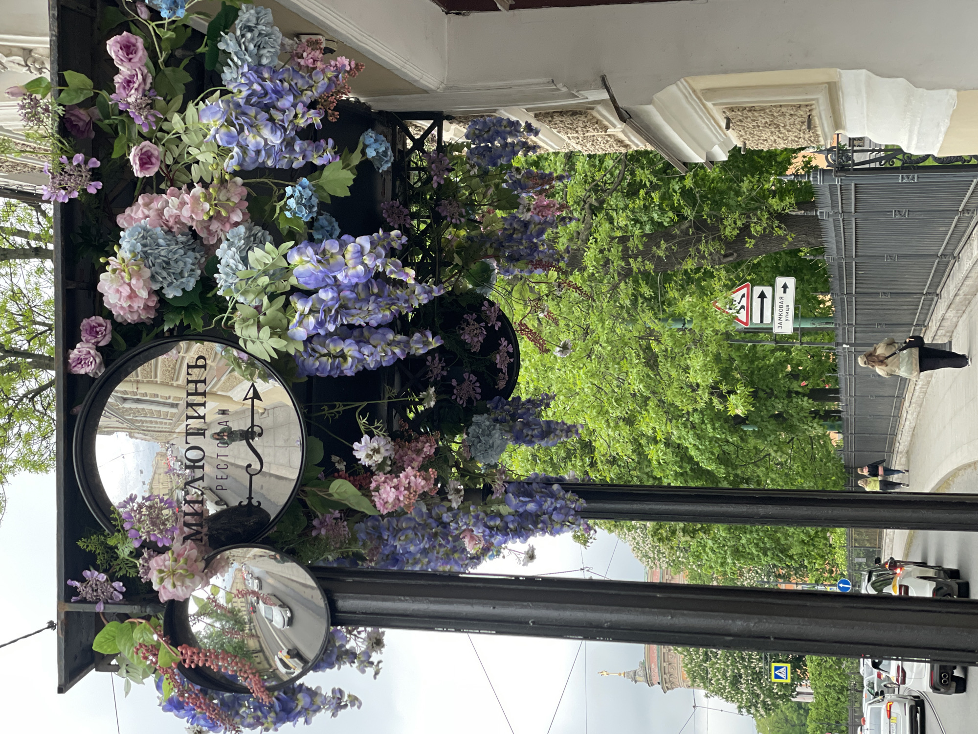 The image shows a street in Saint Petersburg, Russia. A black metal sign with two circular mirrors is decorated with purple and pink flowers. The top mirror displays the reflection of a building with the text "МИЛЮТИНЪ РЕСТОРАН" (Milyutin Restaurant). The bottom mirror reflects a street with cars. A street sign indicating directions, trees, and people walking on the sidewalk are also visible. A building with a spire is visible in the distance.