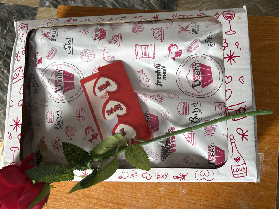 An overhead photograph displays a gift arrangement situated on a light brown wooden surface, with a darker, possibly tiled, surface partially visible in the upper right. The scene is located in Debasco, Nigeria. The central item is a white rectangular cardboard box, approximately 30 cm long by 20 cm wide, featuring a clear plastic window on its lid. The box's exterior is adorned with small red outline illustrations including hearts, stars, bows, a champagne bottle labeled "LOVE," and a spade. Inside the box, a white sheet of paper is visible, patterned with red outline graphics of various food items such as burgers, chips, pizza, cupcakes, shrimp, and grilled chicken, along with text labels like "freshly made" and "SNACK." Prominent branding on this paper includes a pink cupcake logo with "EXQUISITE DEBBY" and contact details: "0906419457 @exquisitedebbycakes exquisitedebby.tictogo." An artificial single-stemmed red rose, with a green stem and green fabric leaves, lies diagonally across the box, its bloom positioned towards the top left. Beneath the rose's stem, a small red card with three white heart shapes, each containing one word ("I," "LOVE," "YOU"), rests atop the patterned paper inside the box. No individuals are depicted in the image.