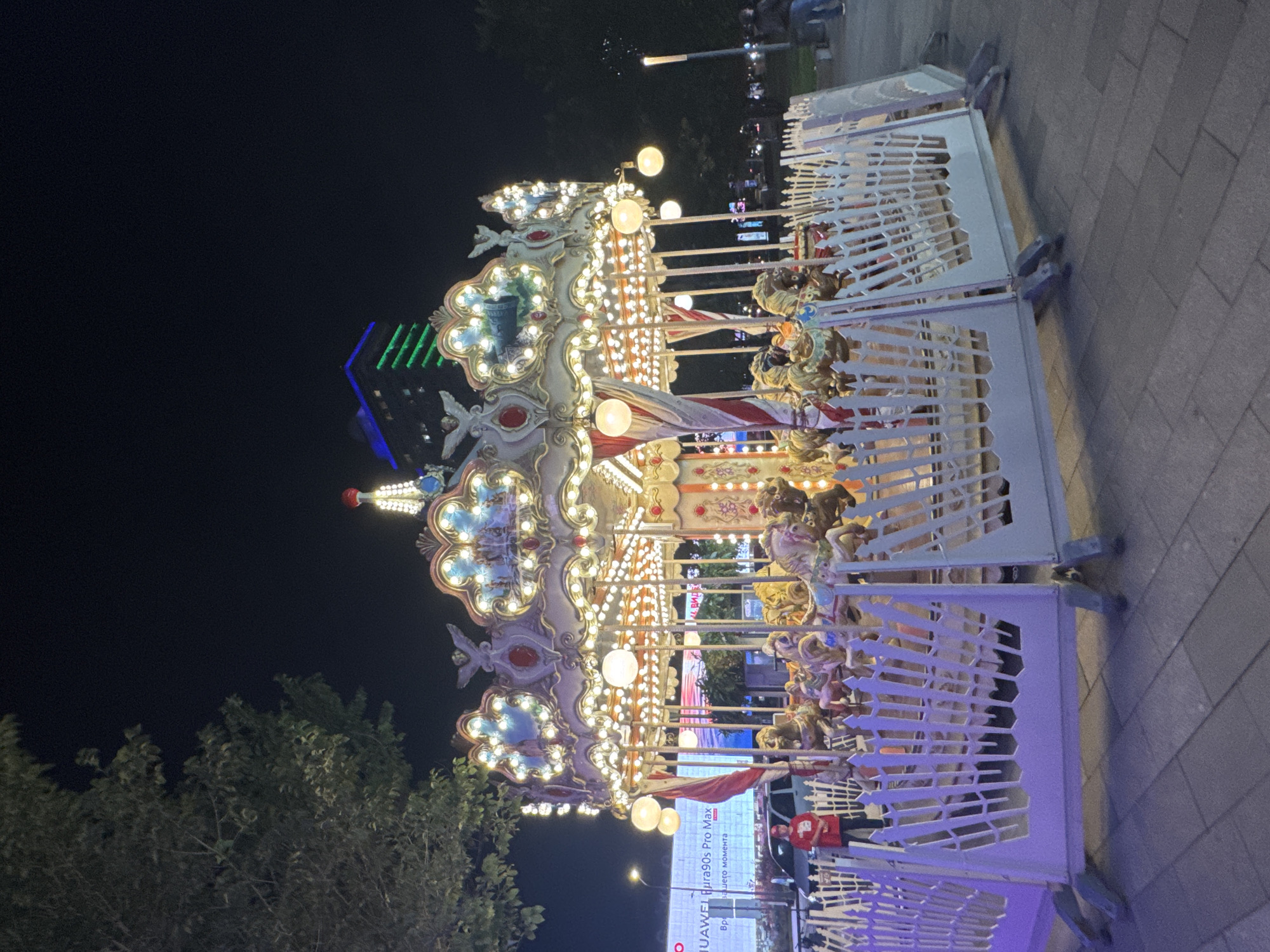 The image shows a carousel at night, likely in a public space within Moscow, Russia. The carousel is brightly lit with numerous bulbs and has an ornate, classic design with figures of horses. It is surrounded by a white, decorative fence. A person in a red shirt and face mask is visible standing near the fence, suggesting an operational amusement. In the background, a modern building with green and blue lights is illuminated against the dark sky, hinting at an urban setting. Trees and streetlights are also present, further contributing to the city environment. The ground appears to be paved.