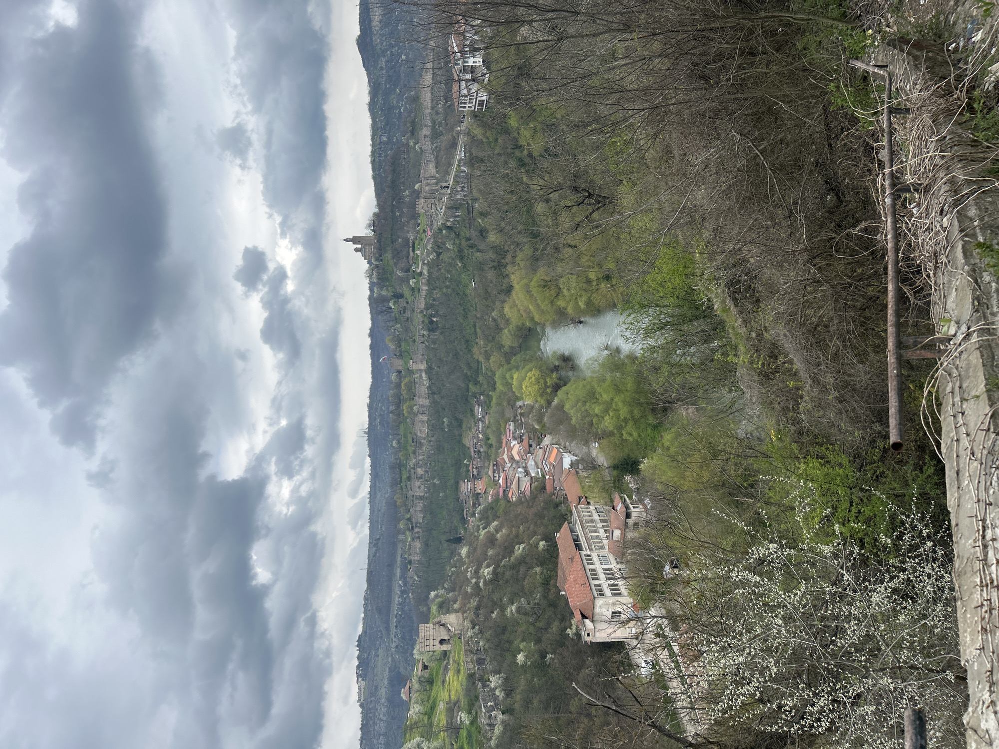 The image depicts a scenic view of Veliko Tarnovo, Bulgaria, taken from an elevated vantage point. The Yantra River winds through a valley lined with lush vegetation. The medieval fortress Tsarevets is visible on the upper left, perched on a hilltop. A lower area hosts houses with red-tiled roofs intermixed with trees. The sky is overcast with dark clouds. In the foreground, a concrete structure and metal pipes are seen.