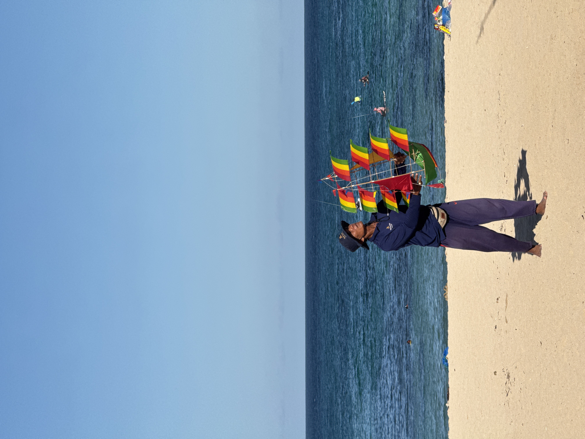 A man stands on a beach in Kuta Selatan, Indonesia, holding a colorful kite in the shape of a sailing ship. He is barefoot and dressed in navy-blue pants and a jacket, and a hat. Behind him, the ocean stretches to the horizon, with a few people visible in the water. To the left of the ocean, the sky is clear and blue. To the right of the man, the sandy beach extends towards the frame's edge. A shadow is cast on the sand near the man's feet.