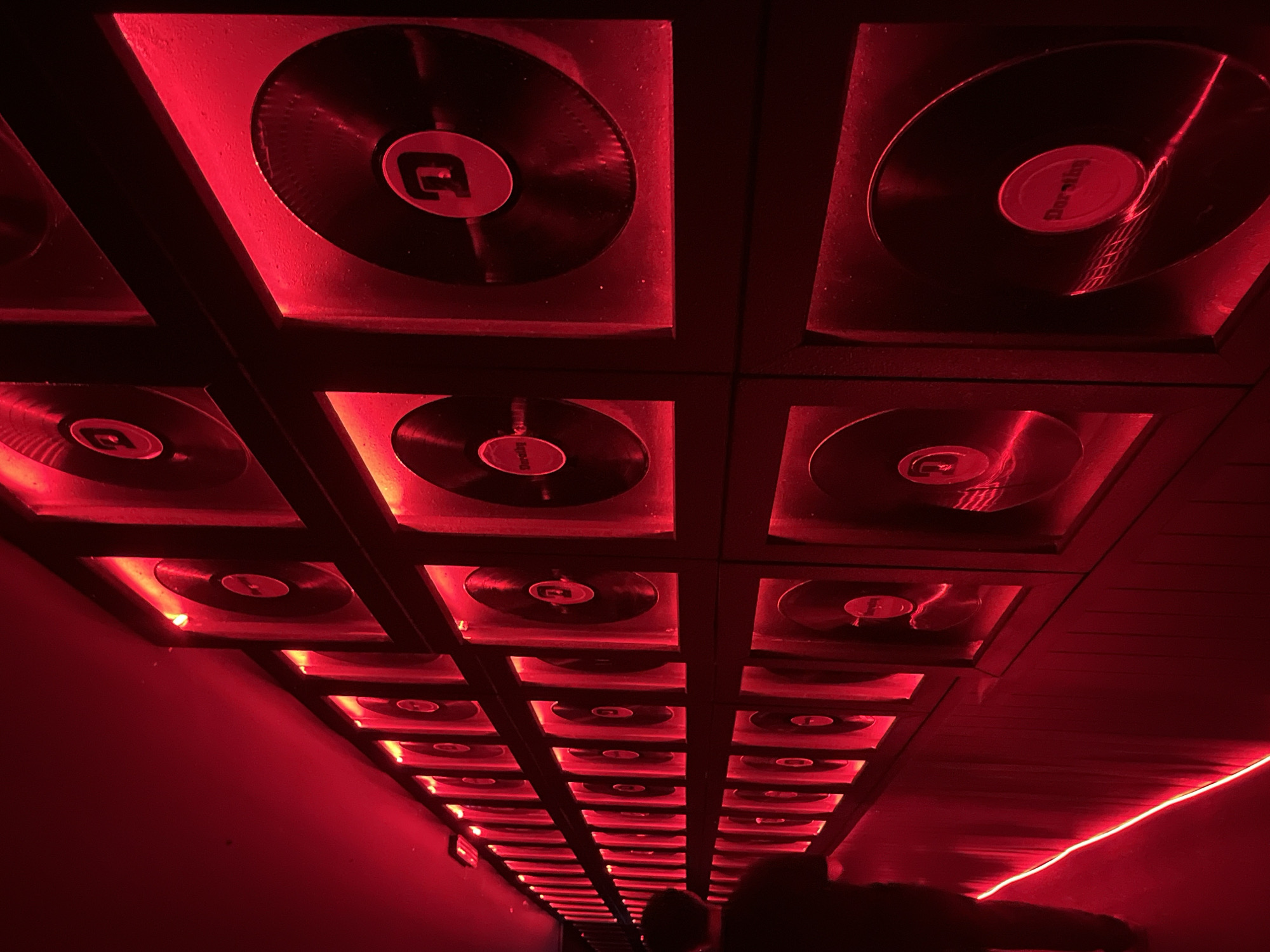 The image captures a red-lit tunnel or hallway with a grid-like ceiling structure, each square containing a vinyl record. The records appear to be illuminated from behind, creating a glowing effect. The overall atmosphere is dark and intensely red. Two people appear at the lower edge of the scene. A thin red strip of light runs along the wall on the right side of the frame. The location is Unknown, Unknown.