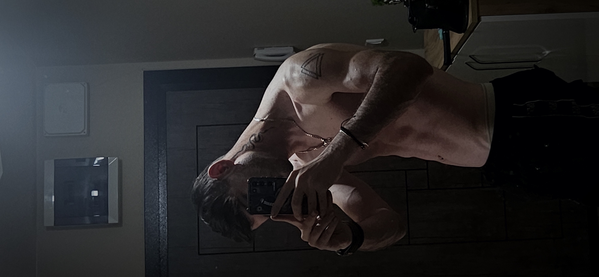 A shirtless man is taking a selfie in a mirror. The image shows his torso and part of his face, with tattoos visible on his shoulder and neck. He is wearing a gold chain. He is holding a phone to take the photo. He is wearing dark shorts or pants. The environment appears to be a bathroom, evidenced by wall-mounted items (mirror, other mounted object) and a door frame visible in the background. The location cannot be determined to be Plovdiv, Bulgaria from the image alone.