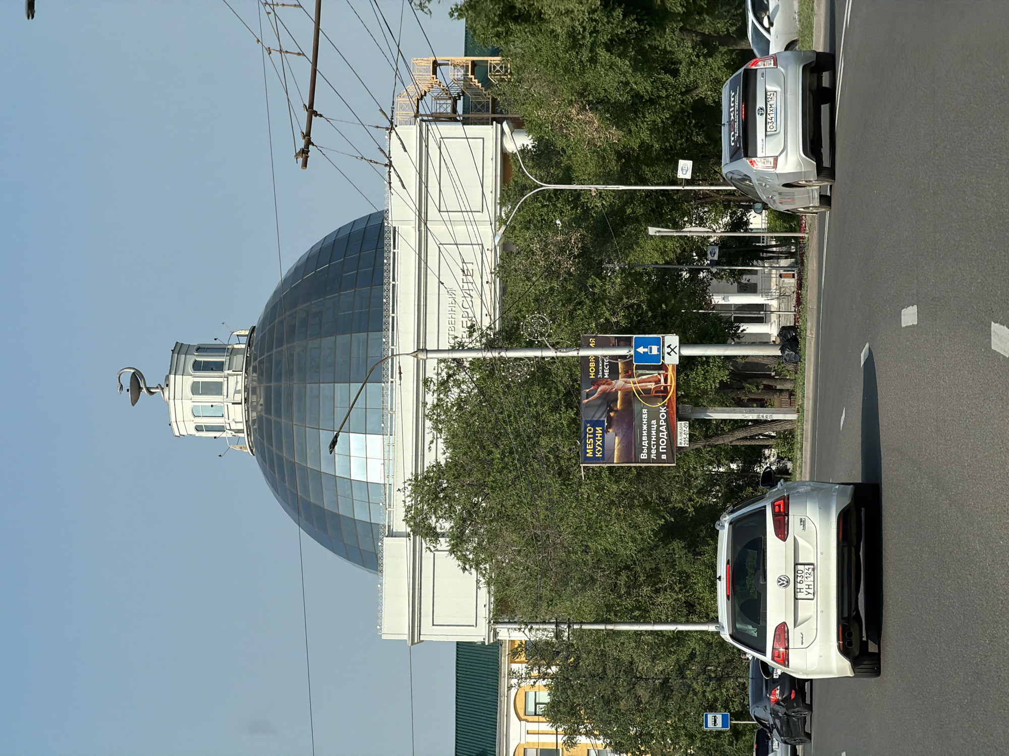 The image captures a street-level view in Krasnoyarsk, Russia, featuring a prominent domed building and traffic. The building, likely an educational institution, has a large, glass-paneled dome topped by a decorative element resembling a stylized bird or phoenix. The facade is white with rectangular indentations. To the left, a yellow building with a green roof is partially visible.
The foreground shows a paved road with white lane markings. Several cars are present: a white Volkswagen Golf station wagon is in the center lane, facing away from the camera. To its left, a black car is partially visible. To the right, a silver hybrid car, possibly a Toyota Prius, is in the right lane, also facing away. A pedestrian crossing sign with an upward arrow and a miner's pickaxe symbol is mounted on a pole.
A billboard advertising "MESTO KUKHNI" (Kitchen Place) and "VYDIVIZHNAYA LESTNITSA V PODAROK" (Extendable ladder as a gift) is positioned to the right of the road, between the cars and the domed building. Overhead wires, likely for an electric trolleybus, crisscross the sky. The scene is set under a clear, light blue sky, indicating daytime. Trees with green foliage line the sides of the road.