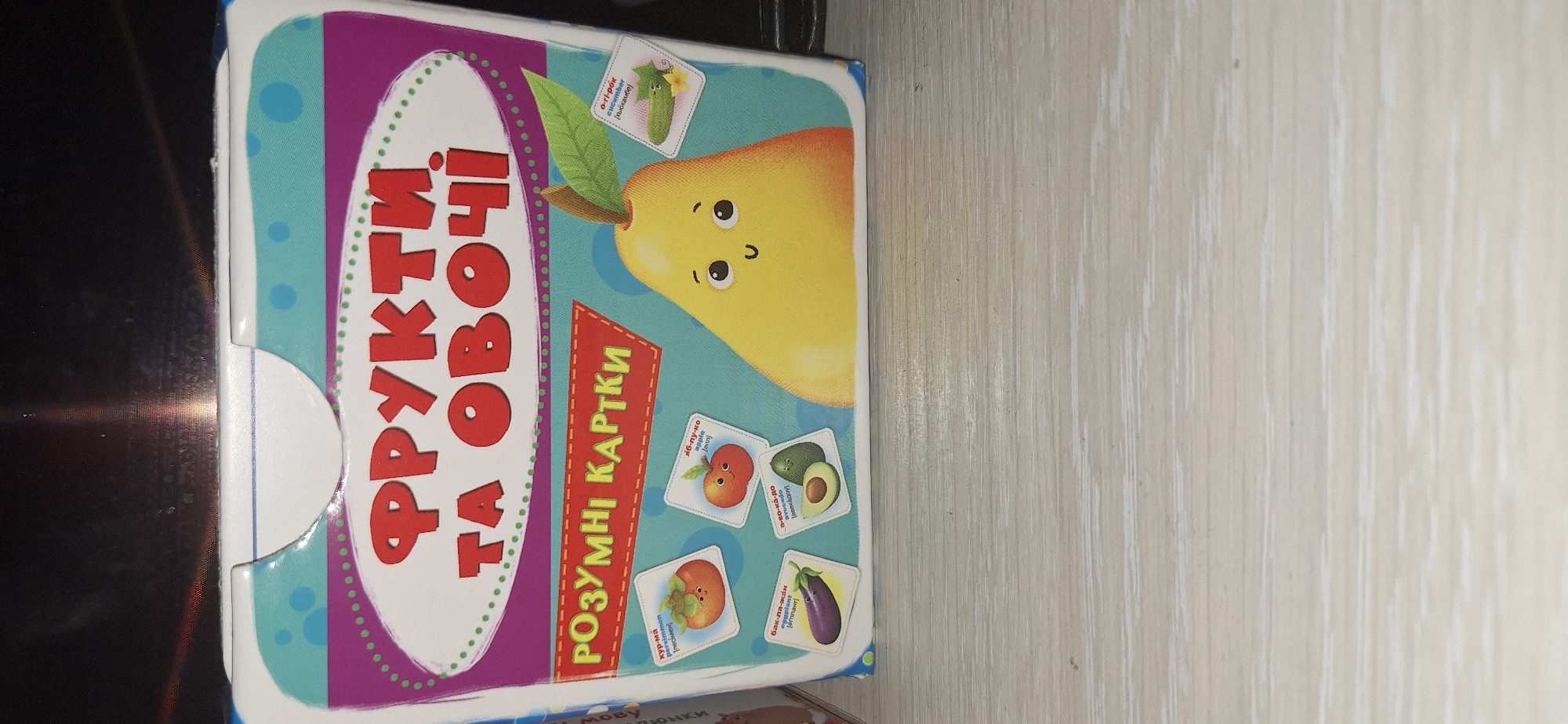 The image shows the front of a children's educational card game box. The box has a colorful design with cartoon fruits and vegetables. The title of the game, "ФРУКТИ ТА ОВОЧІ" (Fruits and Vegetables) and "РОЗУМНІ КАРТКИ" (Smart Cards), is prominently displayed in large red and yellow letters. Several smaller cards featuring individual fruits and vegetables with their names in Ukrainian and English are arranged around a large cartoon pear. The background of the box is a light blue with playful polka dots. The box is placed on a light-colored wooden surface, likely a table or shelf. The scene is well-lit and focused on the product. There is no information or context that would indicate a specific location within Bila Tserkva, Ukraine.