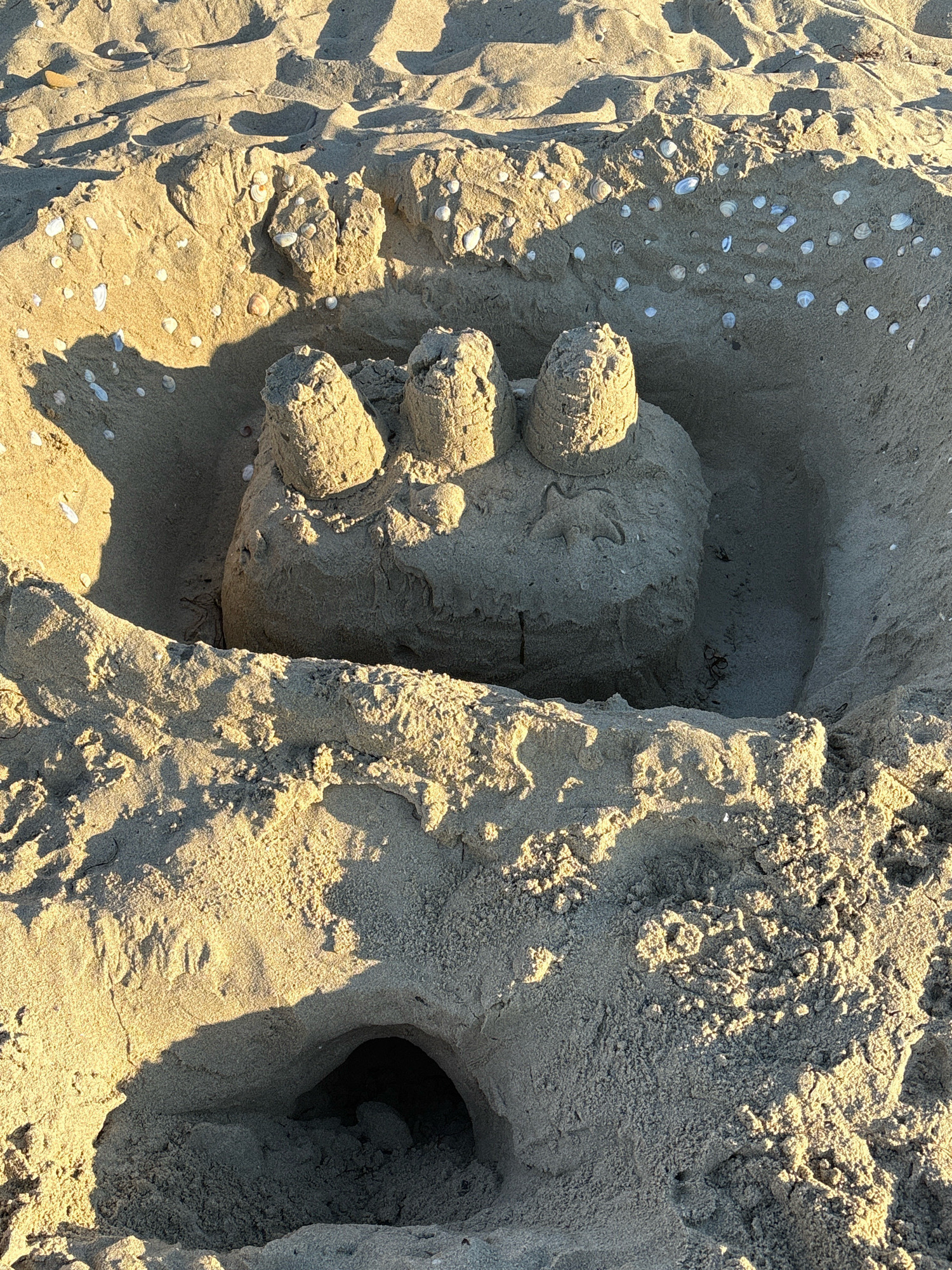 A sandcastle is partially submerged in a shallow pit dug into the sand on a beach. The sandcastle is roughly circular at its base and features three cylindrical turrets on its top. It is decorated with numerous small seashells embedded in its surface. The surrounding sand is textured and marked with shadows cast by sunlight, suggesting it is daytime. The sand also contains some scattered debris, including what appear to be small pieces of seaweed. The environment is a beach, indicated by the sand, shells, and the presence of shadows suggesting open space. The specific location in Sveti Vlas, Bulgaria, is not discernible from the visual information alone.