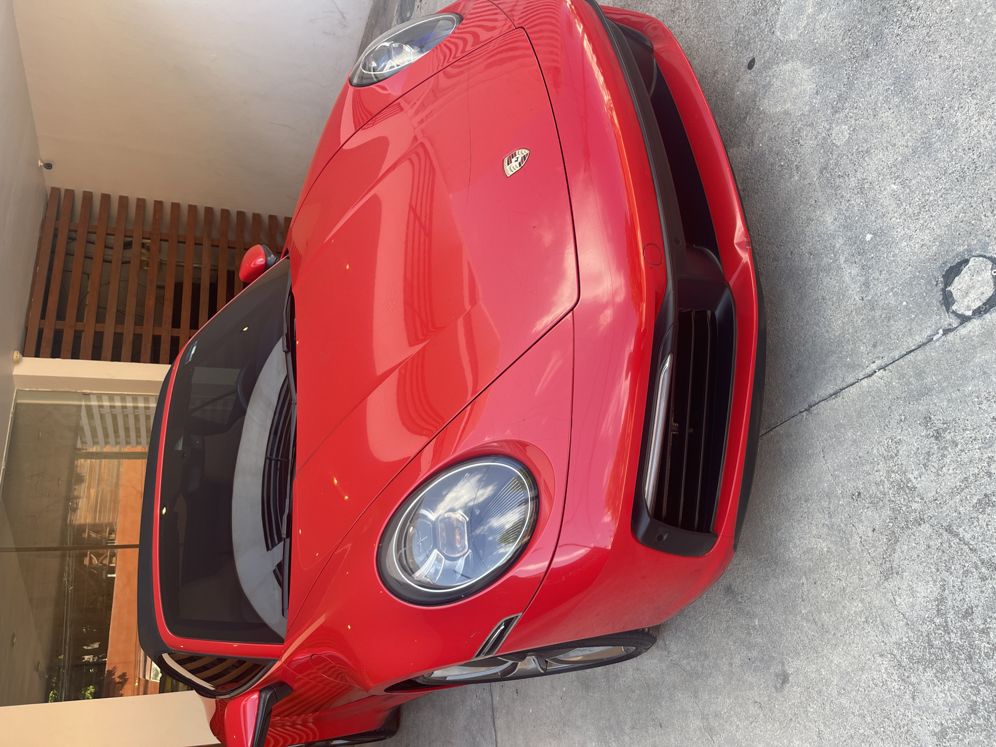 The image is a close-up of a red Porsche 911 parked on a concrete surface. The car's front end is prominent, showcasing its headlights, hood, and front bumper. The Porsche badge is visible on the hood. The environment includes a building with wooden slats and glass windows in the background. Based on available data, the location is Playa del Carmen, Mexico. No people are visible in the image.
