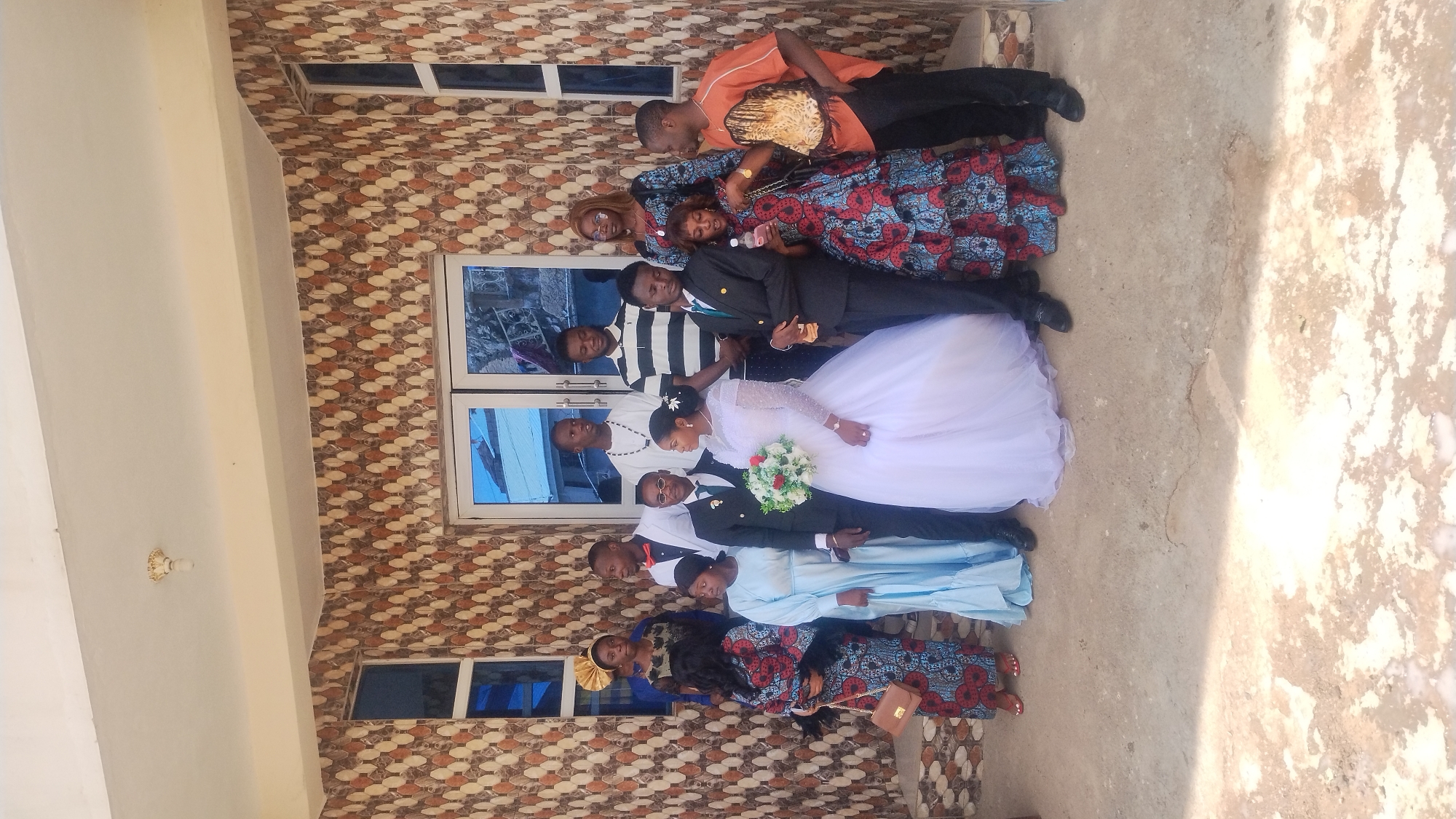 A group of twelve people, likely at a wedding, pose for a photograph in front of a building with tiled walls and glass doors. The building is located in the city of Kabong, Nigeria. The group consists of eight men and four women. The central figures are a bride in a white wedding dress and a groom in a black suit, standing next to each other. The bride holds a bouquet of red and white flowers. Other individuals are dressed in various attire, including suits, traditional African clothing, and casual wear. Some are looking at the camera, while others are looking away or interacting with each other. The ground in front of the building is a concrete surface. The overall scene appears to be an outdoor gathering in a residential or public area.