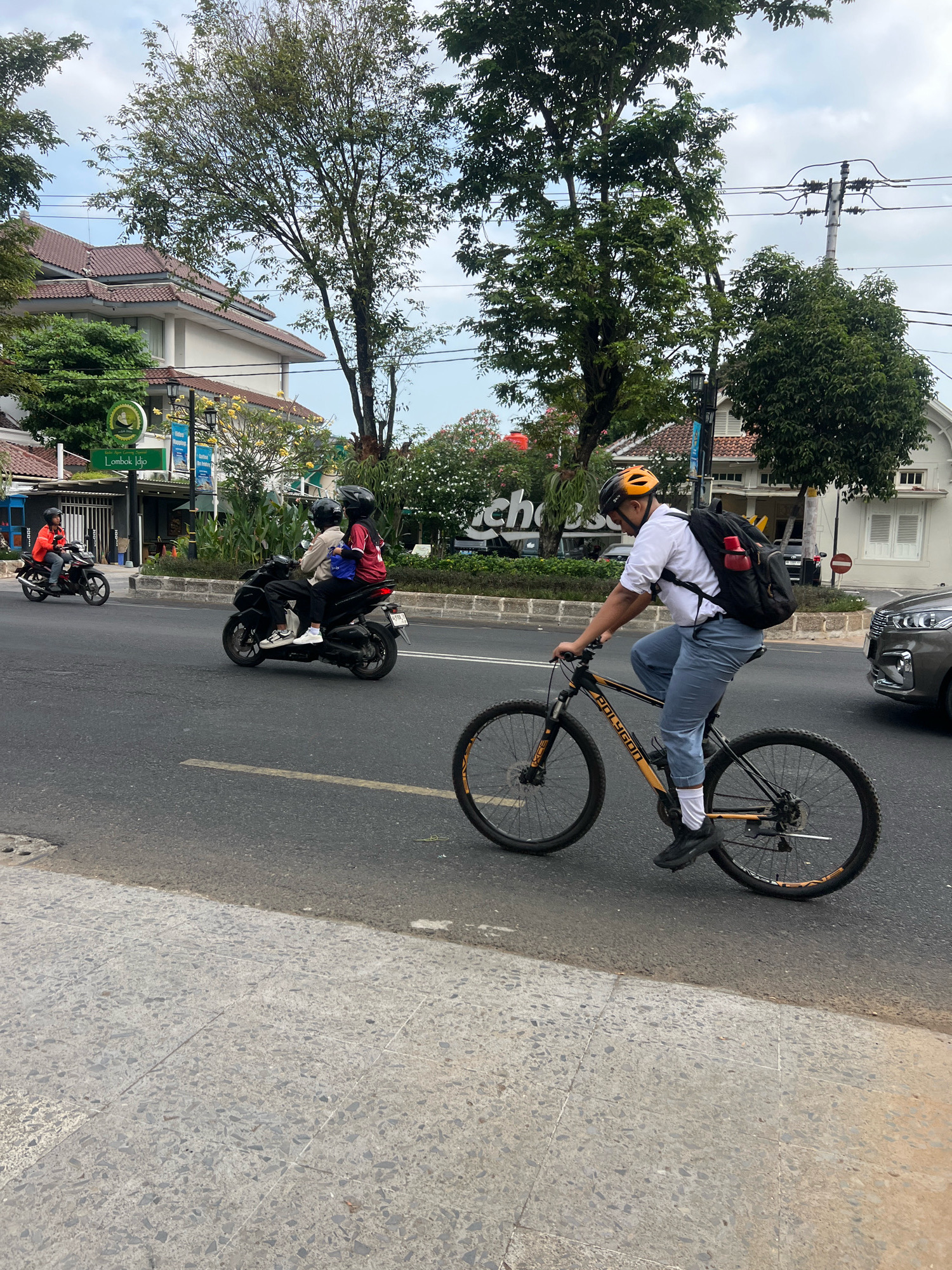 A young male, possibly a student wearing a white collared shirt, blue pants, white socks, and black shoes, is cycling on a paved road in Yogyakarta, Indonesia. He has a yellow helmet on and is carrying a black backpack with a red water bottle attached. The bicycle is black and orange with the brand name "Polygon" visible. The cyclist is in the foreground, with his body angled towards the right side of the frame. In the background, there are trees, buildings with tiled roofs, a street lamp, and an overhead power line. Other vehicles, including two motorcycles with riders, are also visible on the road. One motorcycle, in dark color, is carrying two passengers, both wearing helmets. Another motorcycle in orange jacket is ridden by a single person. A grey car is parked or moving on the right side of the road. The scene appears to be a moderately busy street with a sidewalk in the lower left corner of the image.