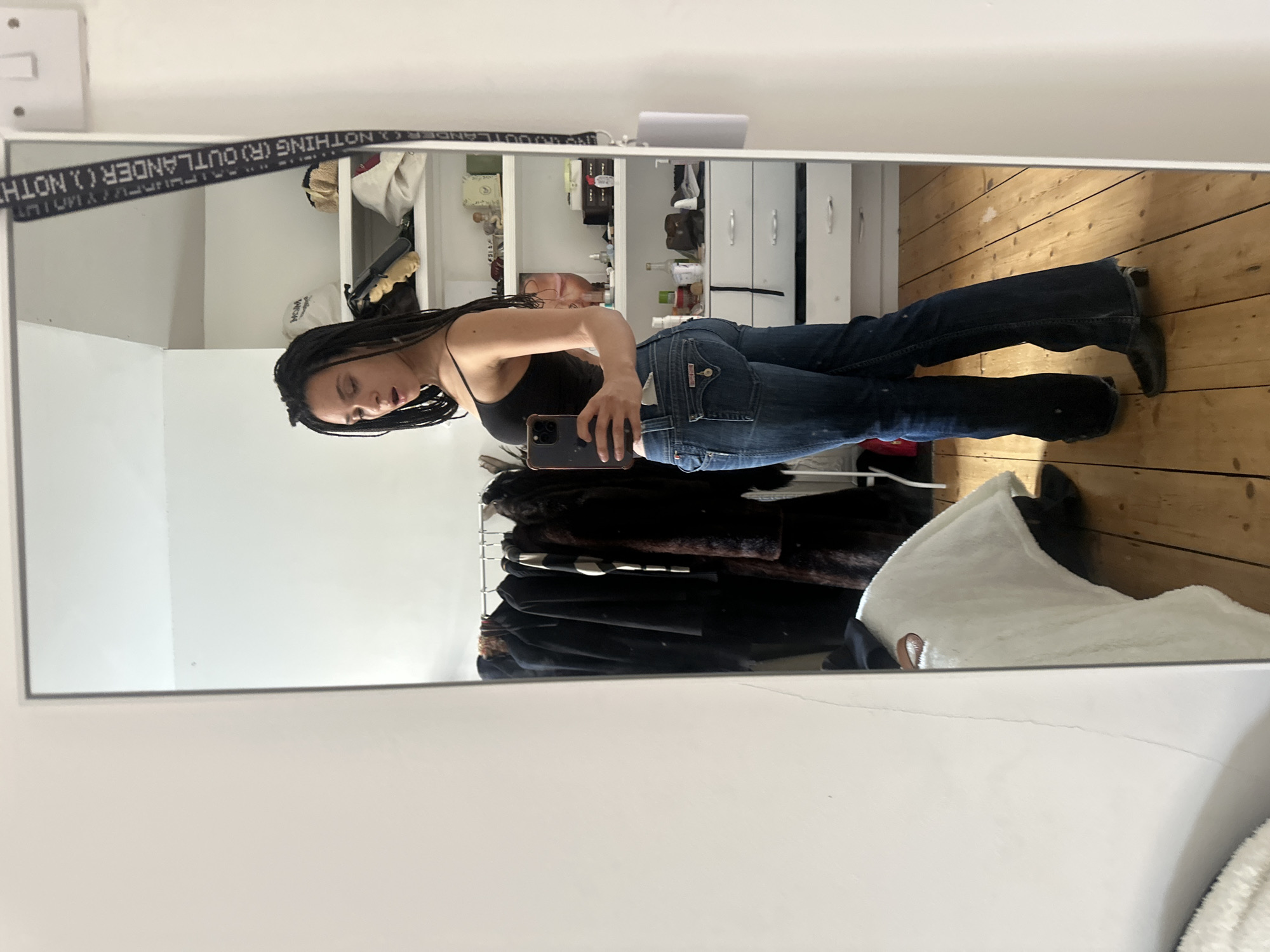 A woman with long dark hair is taking a selfie in a mirror. She is wearing a black tank top and blue jeans. The background includes a wooden floor, a white dresser, and a clothing rack with dark garments. A strap with the text "OUTLANDER (R) NOTHING" is visible along the top of the mirror. The location appears to be indoors, possibly a bedroom. There's no indication this image is in London, United Kingdom.