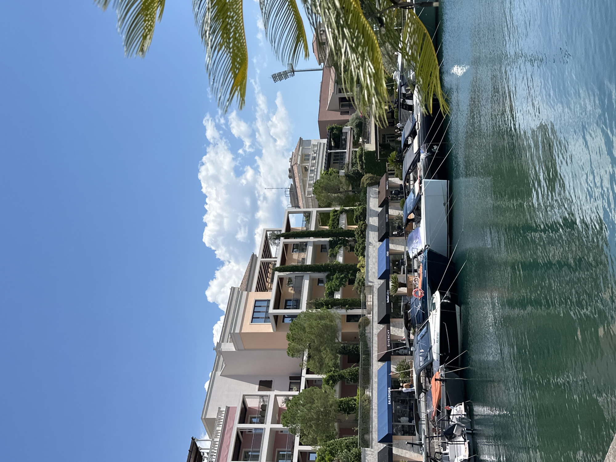 This image depicts a scenic view of a marina in Donja Lastva, Montenegro, under a clear blue sky with scattered white clouds. The foreground is dominated by the calm, dark green water of the marina, with several modern motor yachts docked along the edge. These yachts vary in size and color, with prominent brands like Balenciaga, Givenchy, and Fendi visible on the awnings of the shops lining the waterfront. Behind the yachts, a row of multi-story buildings with balconies and terraces overlooks the marina. These buildings are designed in a contemporary architectural style, featuring a mix of pastel colors and white accents, with lush greenery and tall, slender trees planted around them. In the background, a tall stadium light fixture stands out against the sky, suggesting a nearby sports facility. The overall scene conveys a sense of luxury and leisure in a waterfront setting.