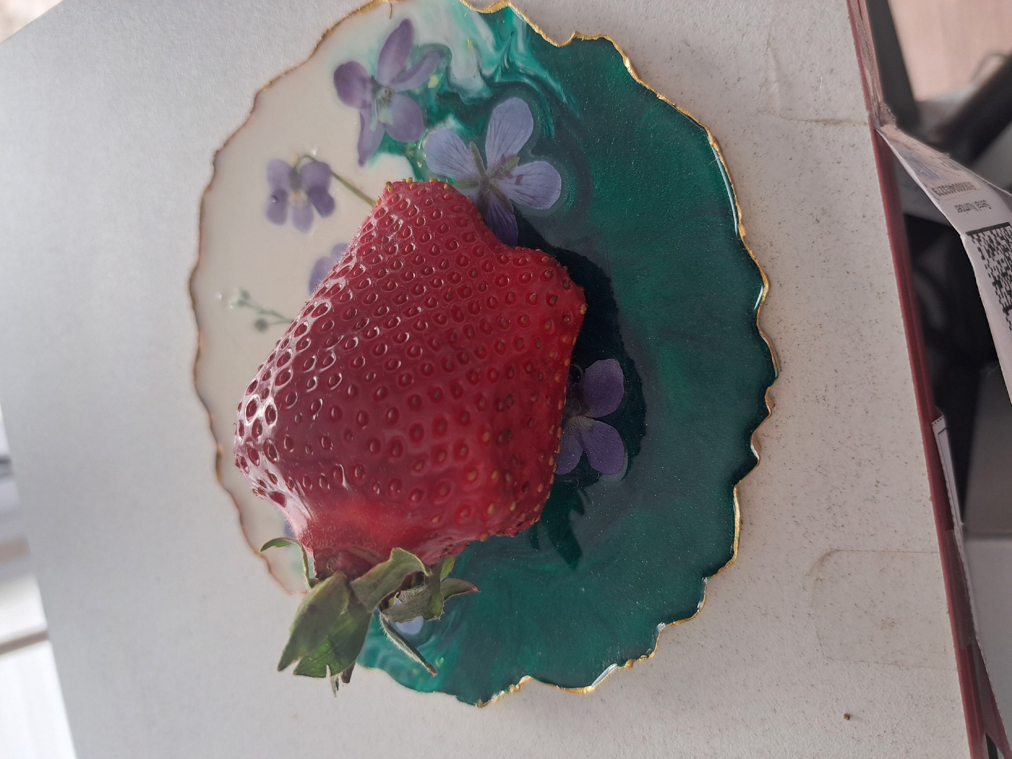 A close-up shot depicts a vibrant red strawberry dominating the frame, positioned atop a round, decorative coaster. The coaster features a two-toned design, with one half showcasing a creamy white background adorned with delicate purple flowers, and the other half displaying a swirled emerald green pattern, also interspersed with purple flowers. A thin gold border traces the edges of the coaster. The scene is set against a pale, textured surface, possibly a wall or tabletop, and a partially visible label is attached to an adjacent object. The location details (Leningradskaya, Russia) are not visually verifiable from the image alone.