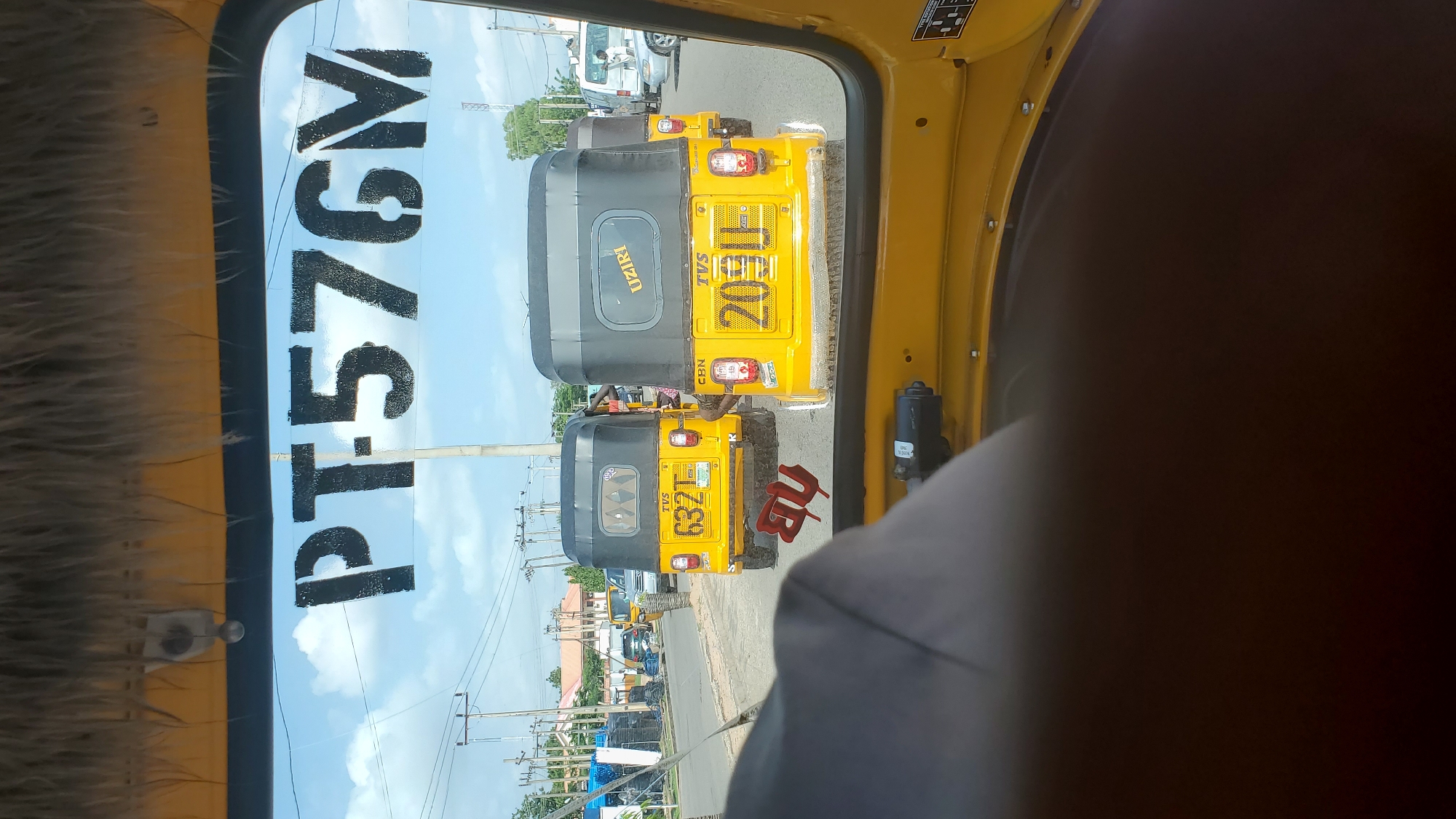 The image is a photograph taken from inside a yellow auto-rickshaw, looking out through the windshield. The windshield has a black decal with white text that reads "PT576V" vertically on the left side. Outside, three auto-rickshaws are visible on a road, apparently stopped in traffic.
The auto-rickshaw directly in front has a gray body and a black roof, with "UZIRI" written on the back. The auto-rickshaw in the middle is yellow with a black roof and has the license plate "20311" on the back. To the right of this, a red symbol is visible on the yellow auto-rickshaw. The auto-rickshaw in the foreground of the road traffic is also yellow with a black roof, displaying the license plate "6321". Further down the road, other vehicles and buildings can be seen, as well as power lines, suggesting an urban or semi-urban environment. The sky is blue with some white clouds, indicating it is daytime and the weather is clear. The overall scene appears to be a typical street scene in Jalingo, Nigeria, with traffic congestion. The photo has a slightly overexposed quality, possibly due to direct sunlight.