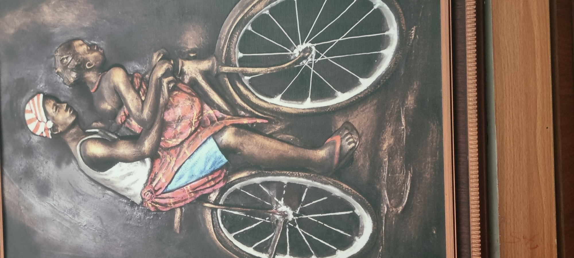 The image depicts a framed artwork of three dark-skinned individuals on a bicycle. One individual wears a white tank top and striped head covering and appears to be pedaling the bicycle. Another individual is behind them, and a third is perched above the handlebar, with the bicycles wheels visible. The artwork is displayed with a wooden frame, with an inner portion of the frame that is dark with a lighter section in the background. The location of Lekki, Nigeria is not able to be confirmed or denied as this is not able to be discerned from the image.