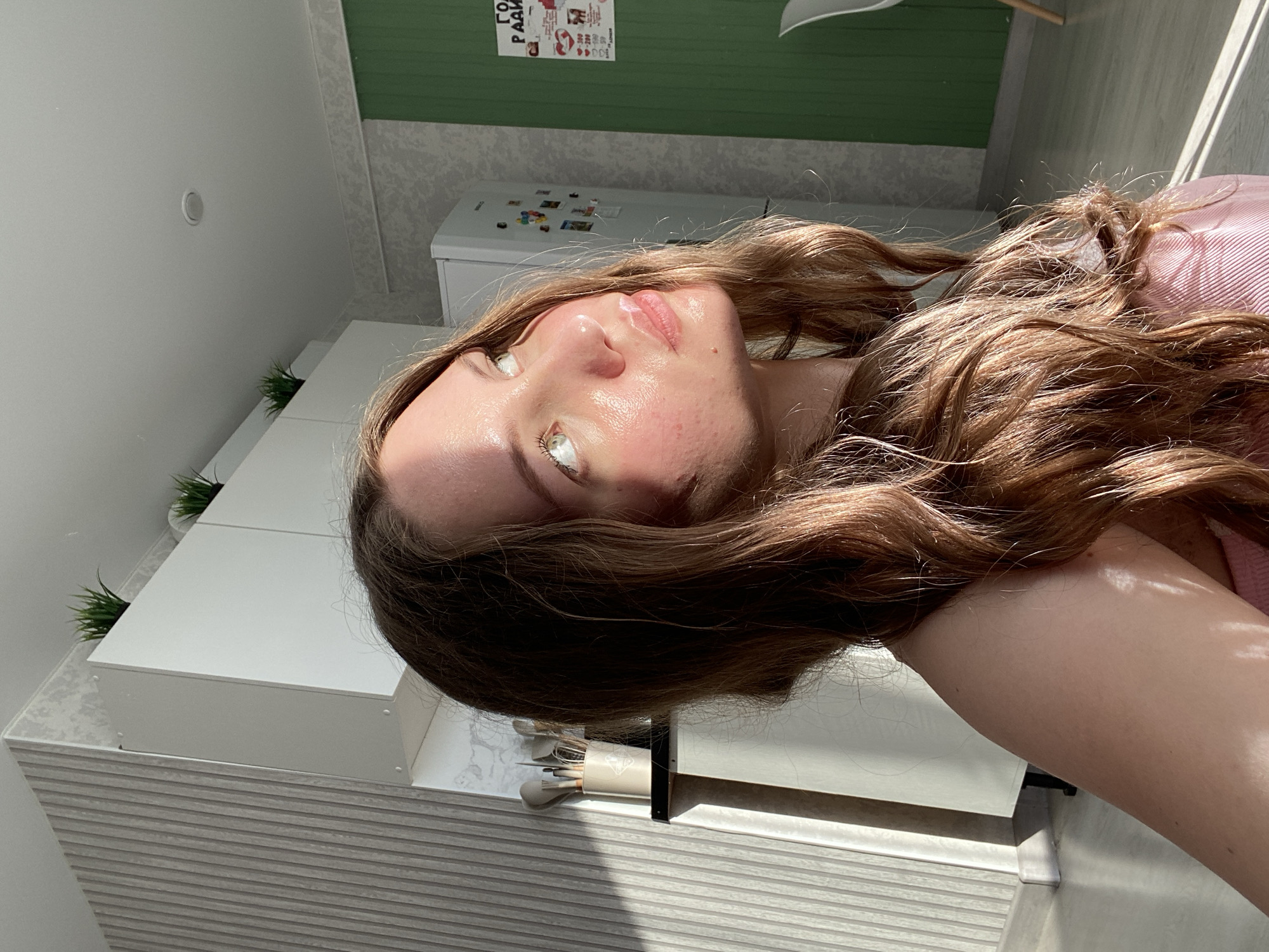 A young woman is captured in a close-up shot, her head tilted back and her face illuminated by a strong light source, likely sunlight. She has long, wavy brown hair that frames her face and spills over her shoulder. Her skin appears to have a dewy sheen, possibly from skincare products or sweat, and shows some redness around her cheeks and nose. Her eyes are open and looking upwards, reflecting the light. She is wearing a pink top.
The background is an interior setting, suggesting a domestic environment. To the left, there are white cabinets, possibly in a kitchen or bathroom, and a glimpse of a refrigerator with colorful magnets on its door. A poster with text in Russian is visible on a green wall in the upper part of the image, accompanied by some small graphics. The lighting creates dramatic shadows on the woman's face and the surfaces around her, indicating it is likely daytime.
The overall mood seems relaxed and perhaps even playful, given the casual pose and natural lighting. The location context is Izhevsk, Russia.