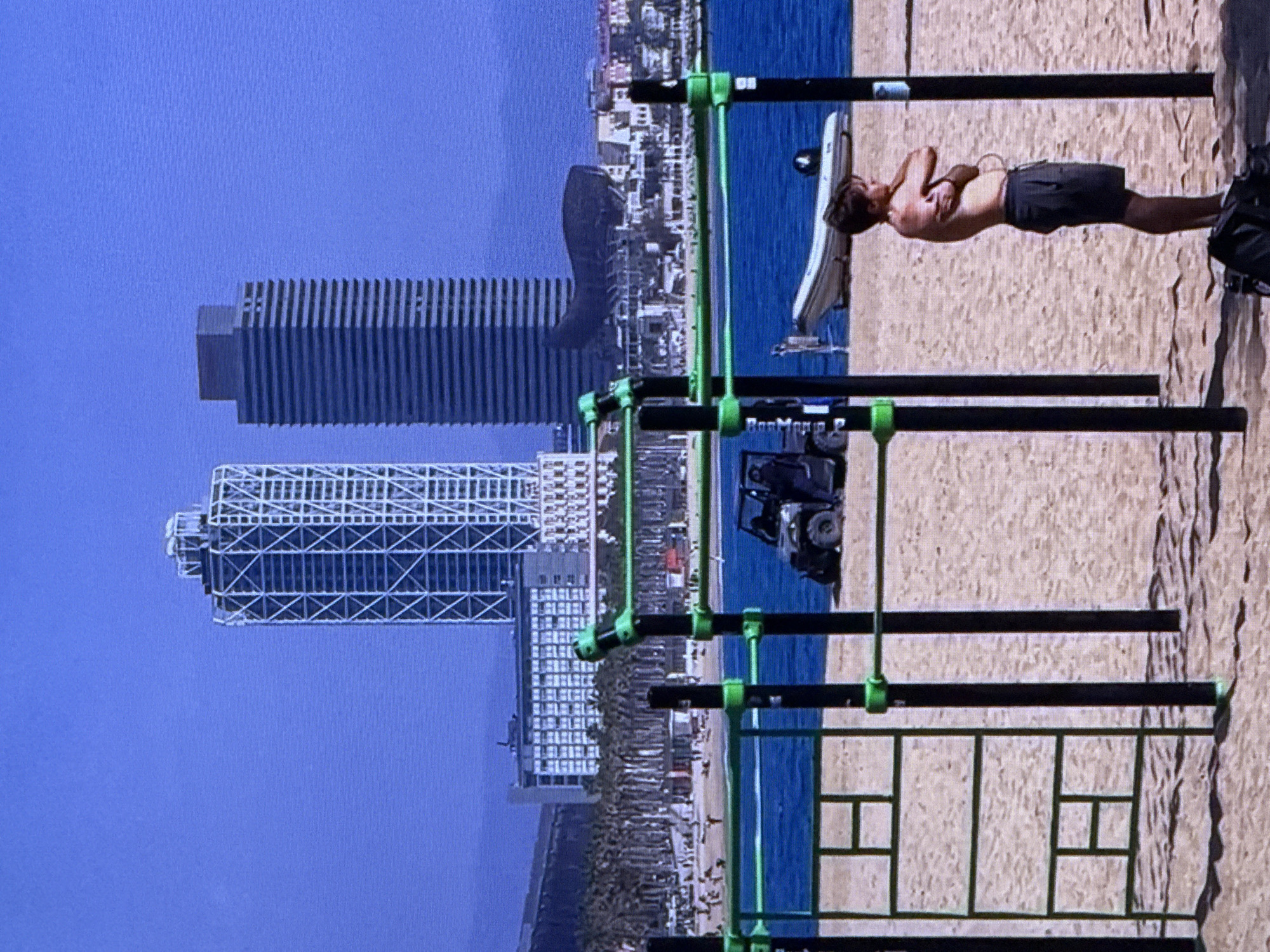 The image depicts a beach scene with a skyline of modern skyscrapers in the background. In the foreground, there is an outdoor fitness area with black poles and green bars. A man is standing on the sand to the right of the frame, facing left, with his arms crossed and his head turned away from the camera. He is wearing dark shorts. Behind him, a small white boat is visible on the blue water, and further back, a black all-terrain vehicle is parked on the sand. Palm trees line the beach in the mid-ground, and a large building complex is situated behind them. The sky is a clear, bright blue. The scene is set in Dubai, United Arab Emirates, as evidenced by the distinctive architecture of the skyscrapers.
