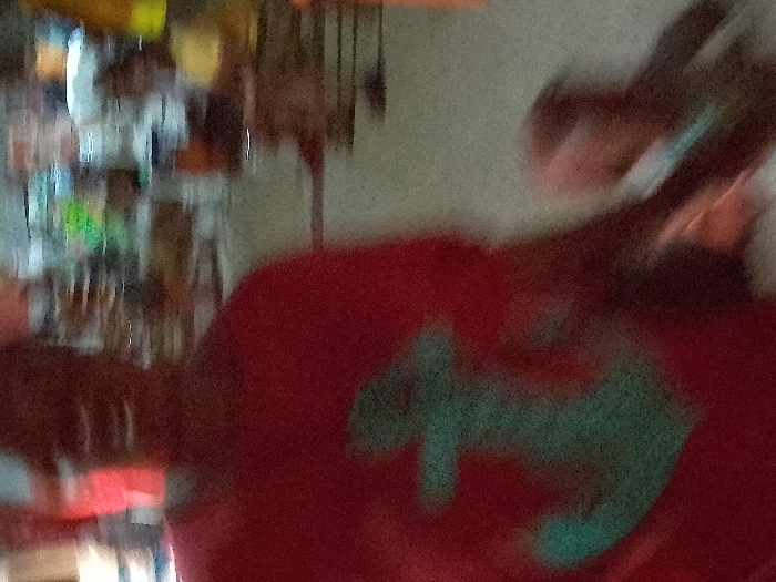 The image depicts a heavily blurred scene within what appears to be an indoor market or shop in Orie Uratta, Nigeria. An adult individual is prominently featured in the foreground, wearing a red short-sleeved shirt with a light blue or teal anchor emblem on the chest. The adult's facial features are indistinguishable due to motion blur. To the adult's right, a child figure is partially visible, also obscured by blur, positioned as if walking alongside the adult and potentially holding their hand. The child is dressed in a light-colored top. The background contains multiple indistinct figures and blurred structures consistent with market stalls or shelves stocked with various items. The ground surface is light in color. The image exhibits significant motion blur, suggesting movement, and indicates low light conditions.