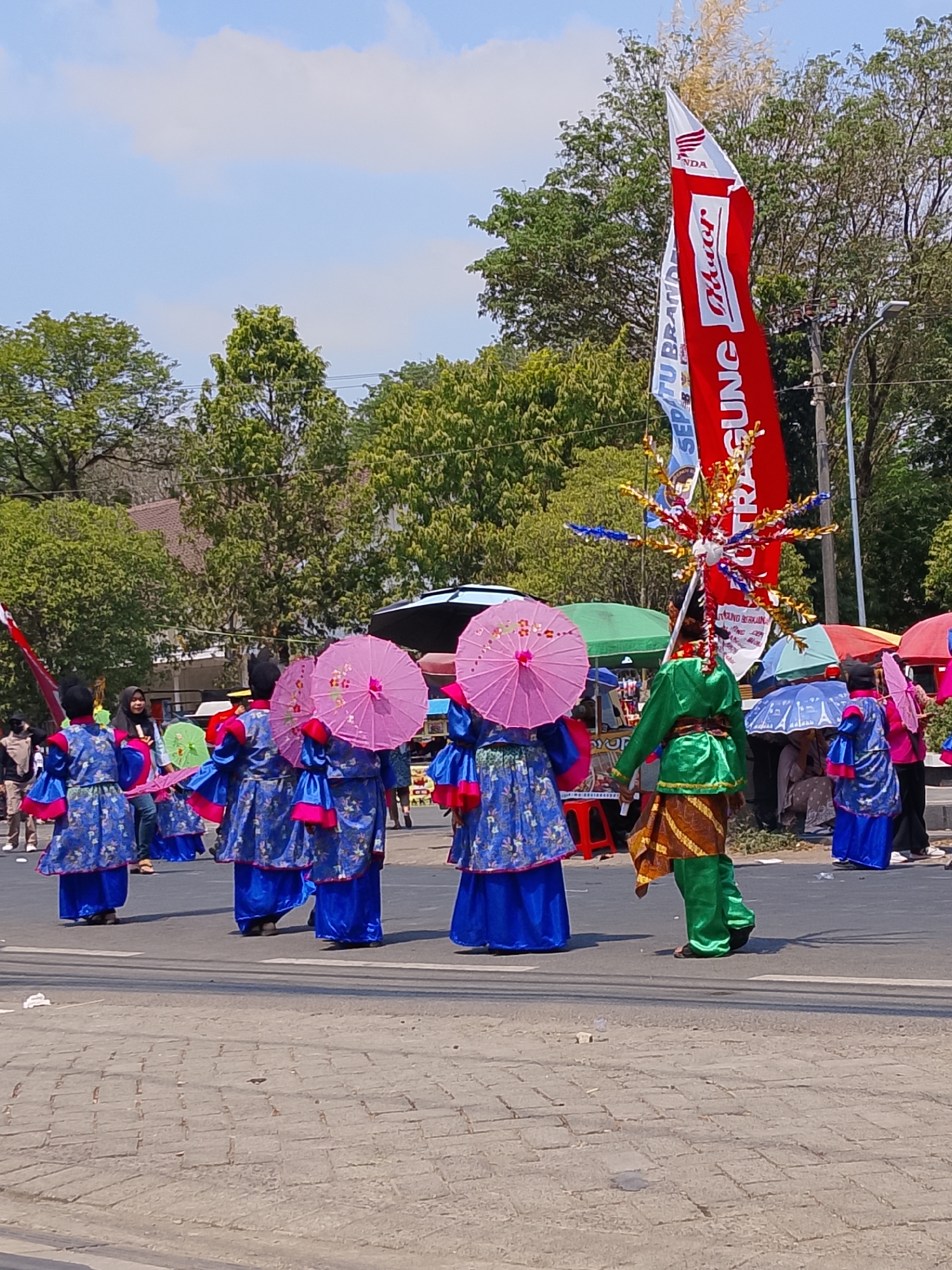 A group of people in traditional blue and pink costumes are walking in a street parade in Balun, Indonesia. The individuals are holding pink parasols and some are wearing hats. There are flags and banners in the background, including a red and white flag with writing on it. The scene appears to be outdoors on a sunny day with trees and buildings visible in the distance. Some spectators are also present, some under umbrellas.