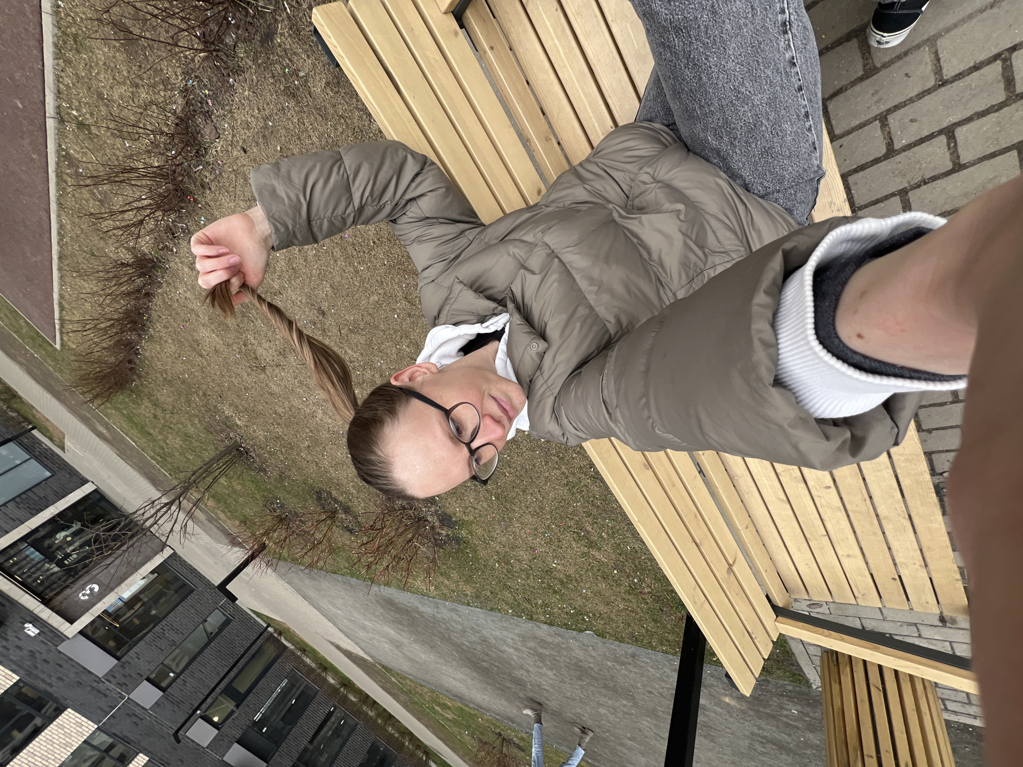 A selfie taken from a high angle shows a woman with blonde hair in a ponytail wearing glasses and a beige puffer jacket sitting on a wooden bench in what appears to be an outdoor area. She's holding her ponytail in her left hand. The bench is made of light-colored wooden slats. The woman is also wearing gray jeans. The surrounding environment includes a building with modern architecture, visible through the windows. There is also a dry grassy area with some bare trees and shrubs, which may be a park or green space. Parts of brick-paved walkways are visible. Although the exact location in Khimki, Russia, cannot be determined from the image alone, the style of buildings and landscaping elements are characteristic of modern urban environments in the city. Another individual is partially visible in the background on the bench.