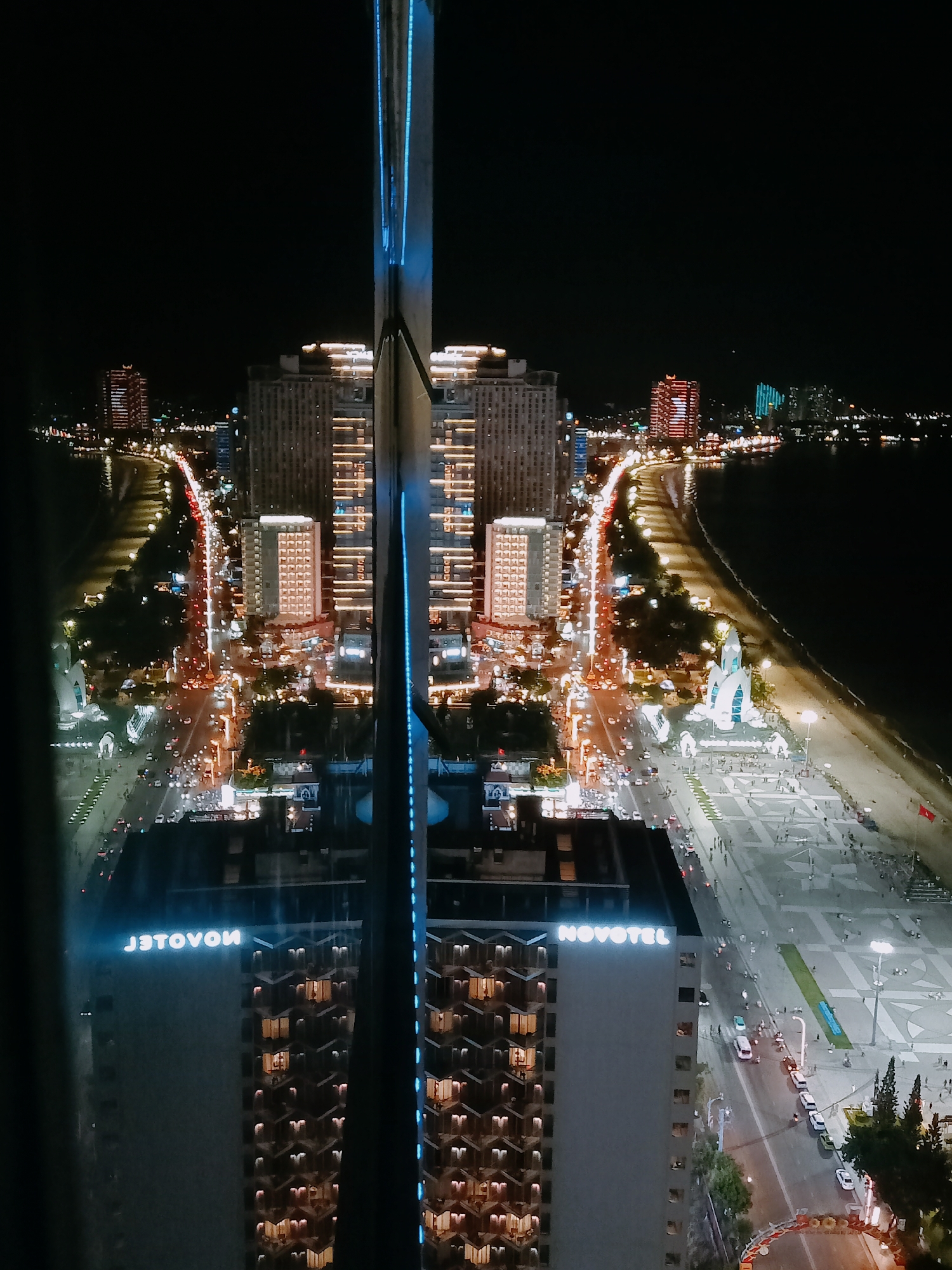 The image shows an aerial nighttime view of Nha Trang Ward, Vietnam, captured through a window, with reflections visible on the glass.
The scene is dominated by illuminated buildings, including a building with "NOVOTEL" signage, suggesting a hotel. Additional high-rise structures with lit windows are visible in the distance. The image captures the nightlights of the city, showing an array of illuminated buildings and streets. A coastal area runs along the scene, with a visible road and beachfront.
There is no observable evidence of people. The overall environment is urban and bustling with light activity associated with nighttime in the city. The composition features a vertical element, possibly part of the building from where the photo was taken, which acts as a central divider.