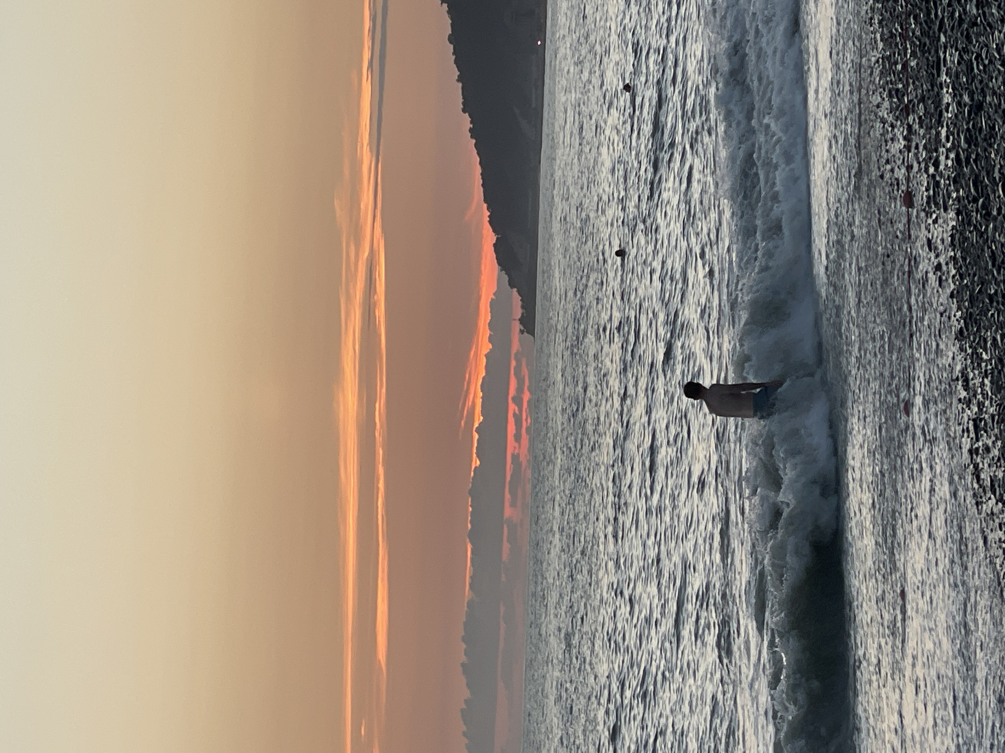 A vertical, eye-level, medium shot captures a person standing in the ocean waves on a beach during sunset. The sky is a gradient of pale yellow at the top, transitioning to shades of orange and pink as it meets the horizon. Thin, wispy clouds are visible, illuminated by the setting sun, with a darker band of clouds lower down. The sea is a deep blue-grey with visible ripples and waves. In the foreground, a person, seen from behind, is partially submerged in the breaking waves, wearing blue shorts. The beach in the lower portion of the image is covered in dark grey and brown pebbles. In the distance, a dark, tree-covered hillside forms the coastline, with a small lighthouse-like structure visible on its edge. The overall scene depicts a serene moment at the end of the day, likely on one of the pebble beaches of Sochi, Russia, given the characteristic coastline.