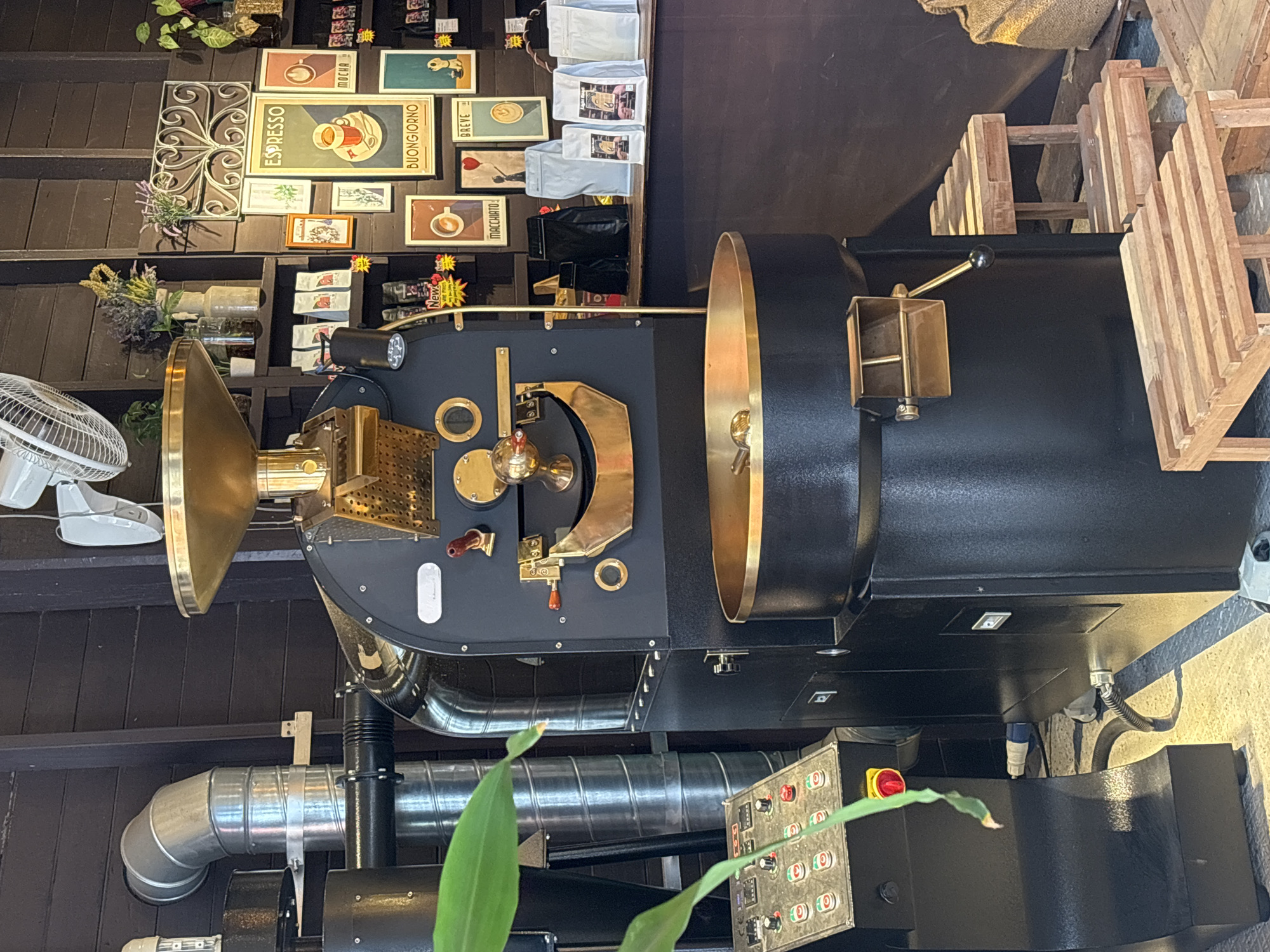 This is a medium shot, eye-level photograph of a commercial coffee roaster in a shop that appears to be in Chiang Mai City Municipality, Thailand. The roaster is a large, black, industrial machine with brass accents, positioned centrally in the frame. To the left of the roaster is a control panel with numerous buttons and a red emergency stop button, and to the right are small wooden stools and a burlap sack. Behind the roaster, a dark wooden wall is decorated with framed pictures and a vase of flowers. Several bags of coffee beans are arranged on a counter behind the roaster. The lighting suggests an indoor commercial environment, possibly a coffee shop or roastery. There are no people visible in the frame.