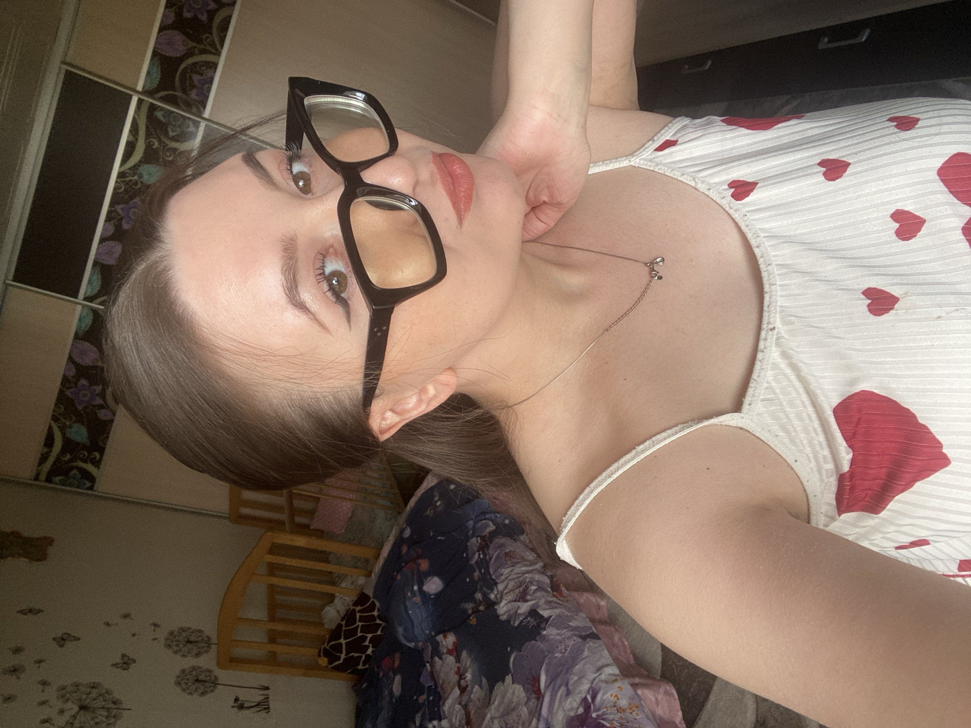 A young woman with dark hair and glasses poses for a close-up selfie. She wears a white top with red heart patterns. The background includes a wardrobe with patterned doors and a child's crib. The wallpaper features dandelion and butterfly patterns. While a city is mentioned, the details in the image do not confirm the location of Chaikovsky, Russia.