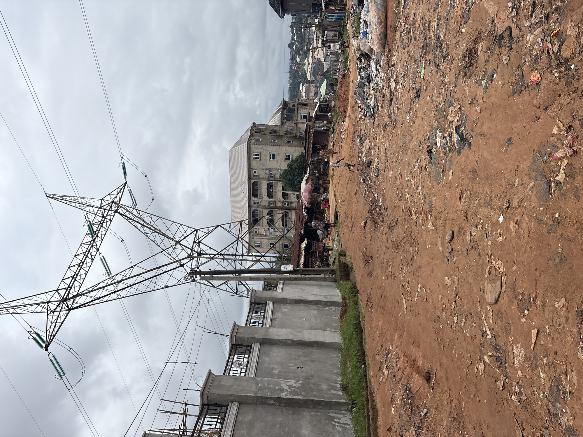In Nkpor, Nigeria, a wide, dusty red dirt road is strewn with garbage, including plastic bags, shredded paper, and discarded materials. In the foreground, the road is heavily littered. A tall, metal electricity pylon looms over the scene, with thick electrical wires crisscrossing the sky, which is overcast. To the left of the road, a tall concrete wall with decorative metalwork extends into the background. Beyond the wall, a large, multi-story building with a light-colored roof and multiple balconies is visible. In the middle ground, a small market appears to be operating under makeshift awnings, with several people browsing or tending to stalls. A child runs across the dirt road. Further into the background, more buildings of various sizes and styles form a dense urban landscape, and more debris is visible along the sides of the road.