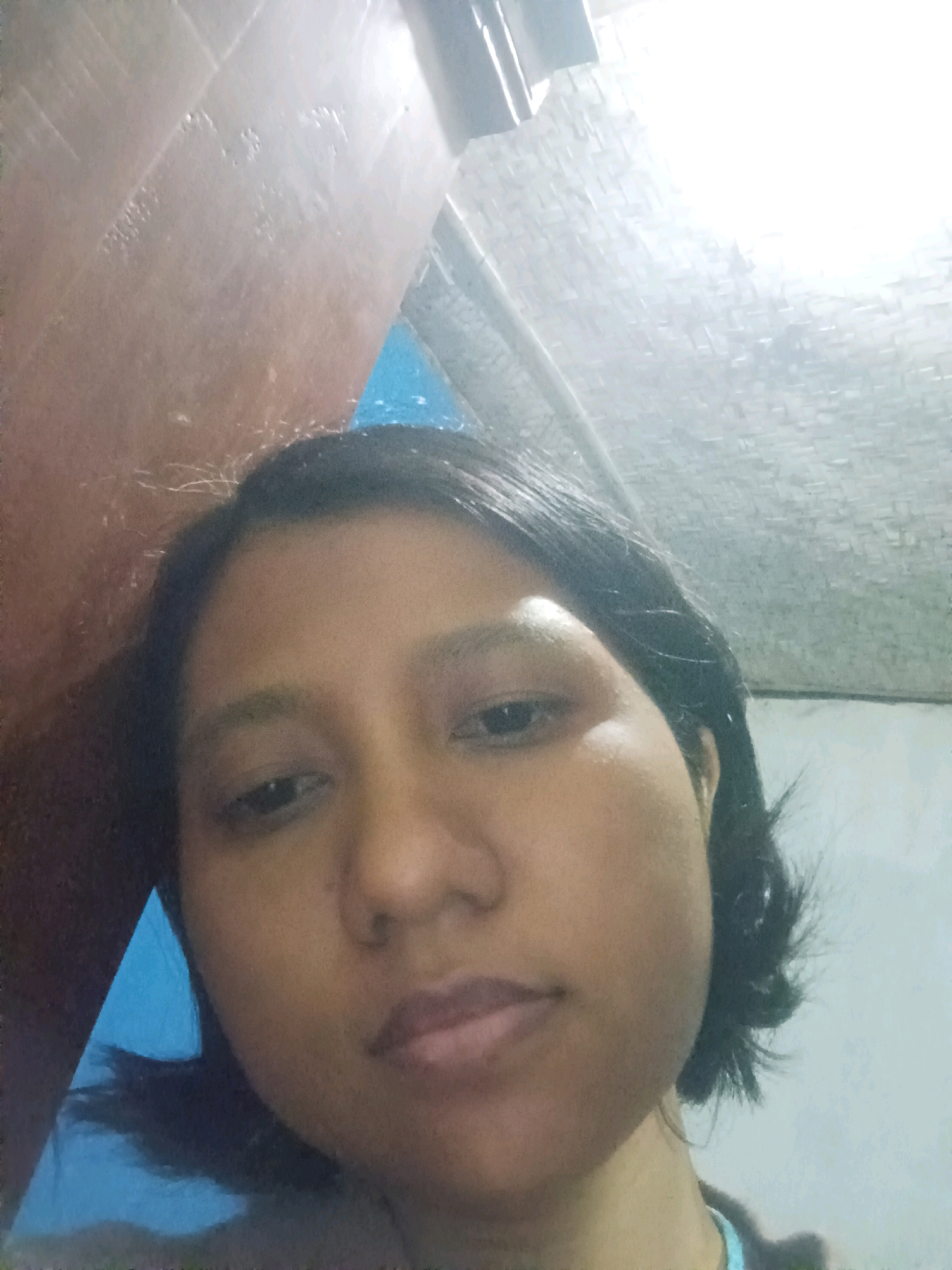 A close-up selfie shows a young woman's face filling most of the frame. She has dark hair and is looking directly at the camera. The background is blurry and appears to be an indoor setting, with a textured, light-colored ceiling visible at the top. To the left, there is a reddish-brown wall, and to the right, a lighter, grayish wall. A triangular piece of bright blue is visible in the upper left quadrant. The lighting is uneven, with a bright light source in the upper right corner creating a glare. There are no discernible city landmarks or environmental clues to suggest a specific location within Canga'an, Indonesia.