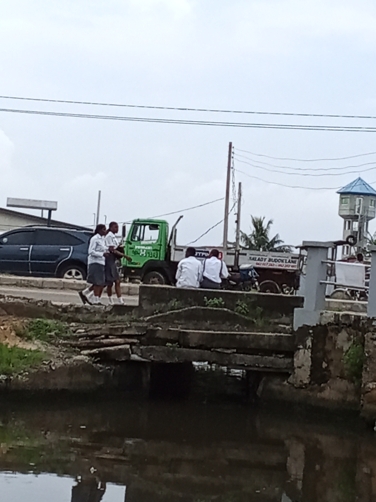 This image depicts an overcast daytime scene in Ado, Nigeria, featuring a waterway or canal in the foreground spanned by a basic concrete culvert or bridge. The water is dark and still, reflecting some light.
In the midground, two young people, likely students, are walking from left to right on a paved area next to a road. They are dressed in white short-sleeved shirts and dark skirts, with white socks. Behind them, a dark blue car is parked on the roadside. Further to the right, two other young people, also appearing to be students in white shirts and dark bottoms, are sitting on the concrete edge of the waterway structure, with their backs mostly to the camera.
The road behind these individuals shows traffic, including a bright green utility or refuse truck and another dark-colored flatbed truck. The flatbed truck has "SALADY BUDDYLANE" and partial phone numbers "080 617 263--" and "080 242 480--" visibly printed on its side. Utility poles with multiple overhead wires stretch across the scene. In the background, a tall water tower with a blue, conical roof is prominent, alongside palm trees and other distant structures, all under a uniformly grey, cloudy sky. The overall impression is an everyday street scene in a semi-urban environment.