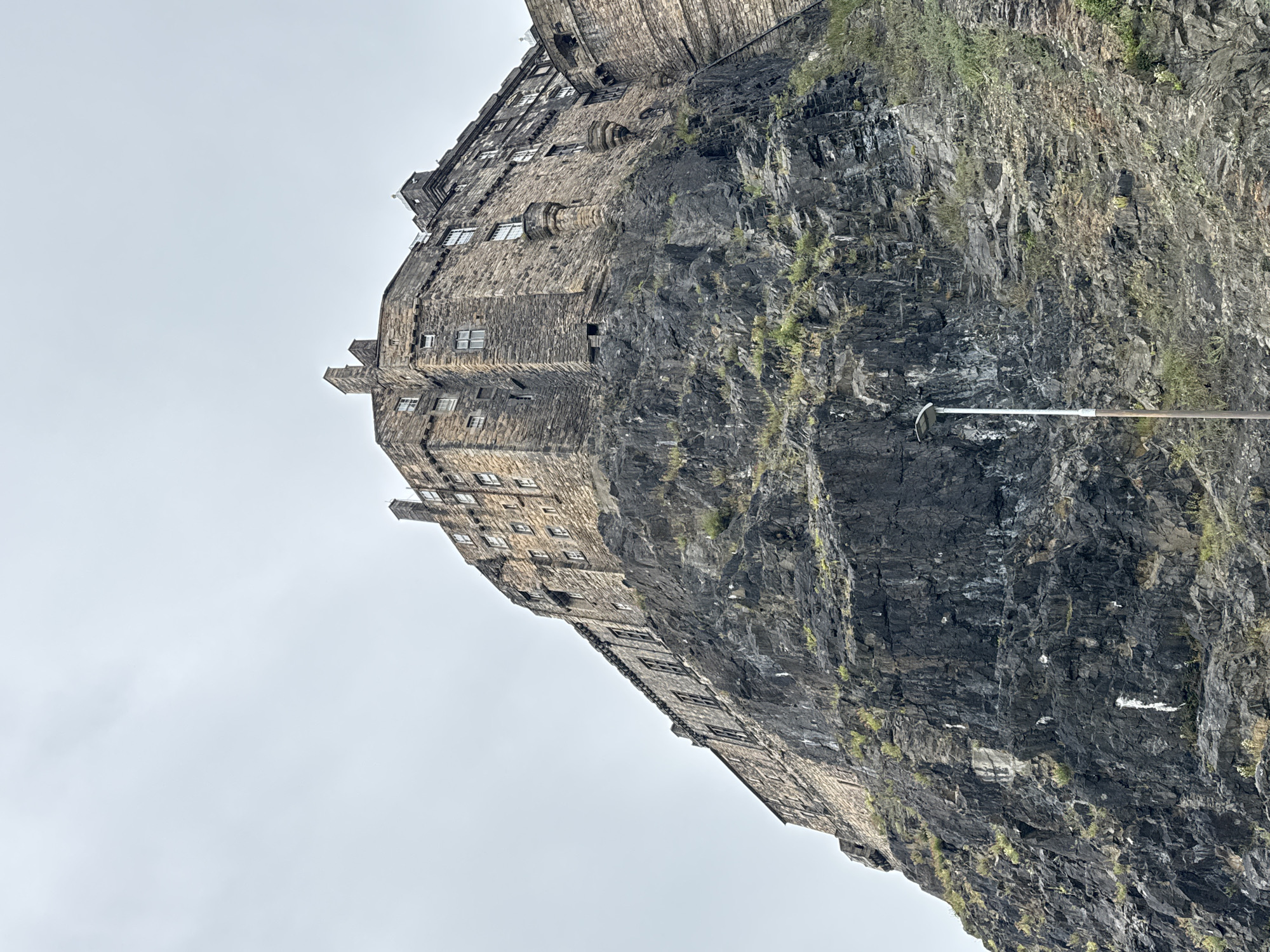 This is a low-angle, eye-level, full shot of Edinburgh Castle, situated on Castle Rock in the City of Edinburgh, United Kingdom. The castle is constructed from dark, rough-hewn stone and features numerous windows and chimneys. It is perched atop a steep, rocky cliff face that dominates the lower half of the frame. The cliff is uneven and textured, with sparse vegetation growing in its crevices. The sky above is overcast and a uniform pale gray. A single, tall, white lamppost stands on the rocky slope in the lower right quadrant of the image, providing a sense of scale. No people are visible in the frame.