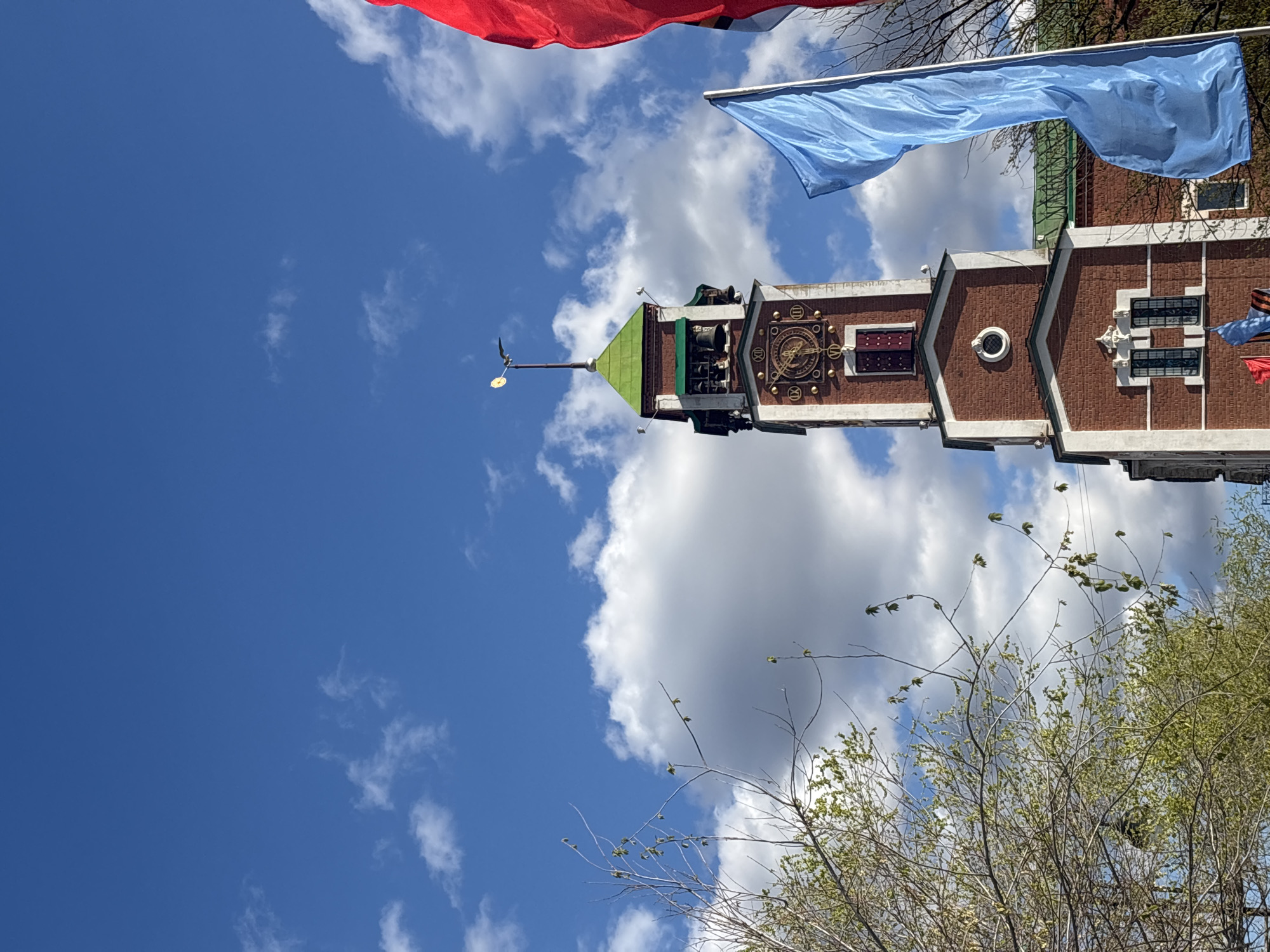 The image captures the clock tower of Orenburg's city administration building against a backdrop of a blue sky dotted with white clouds. A red and a blue flag are visible in the upper part of the frame. The tower features a green, conical roof topped with a weather vane. A clock face with roman numerals is visible on the tower. The tower's architecture includes red brick and white accents. Bare tree branches occupy the lower right portion of the frame.