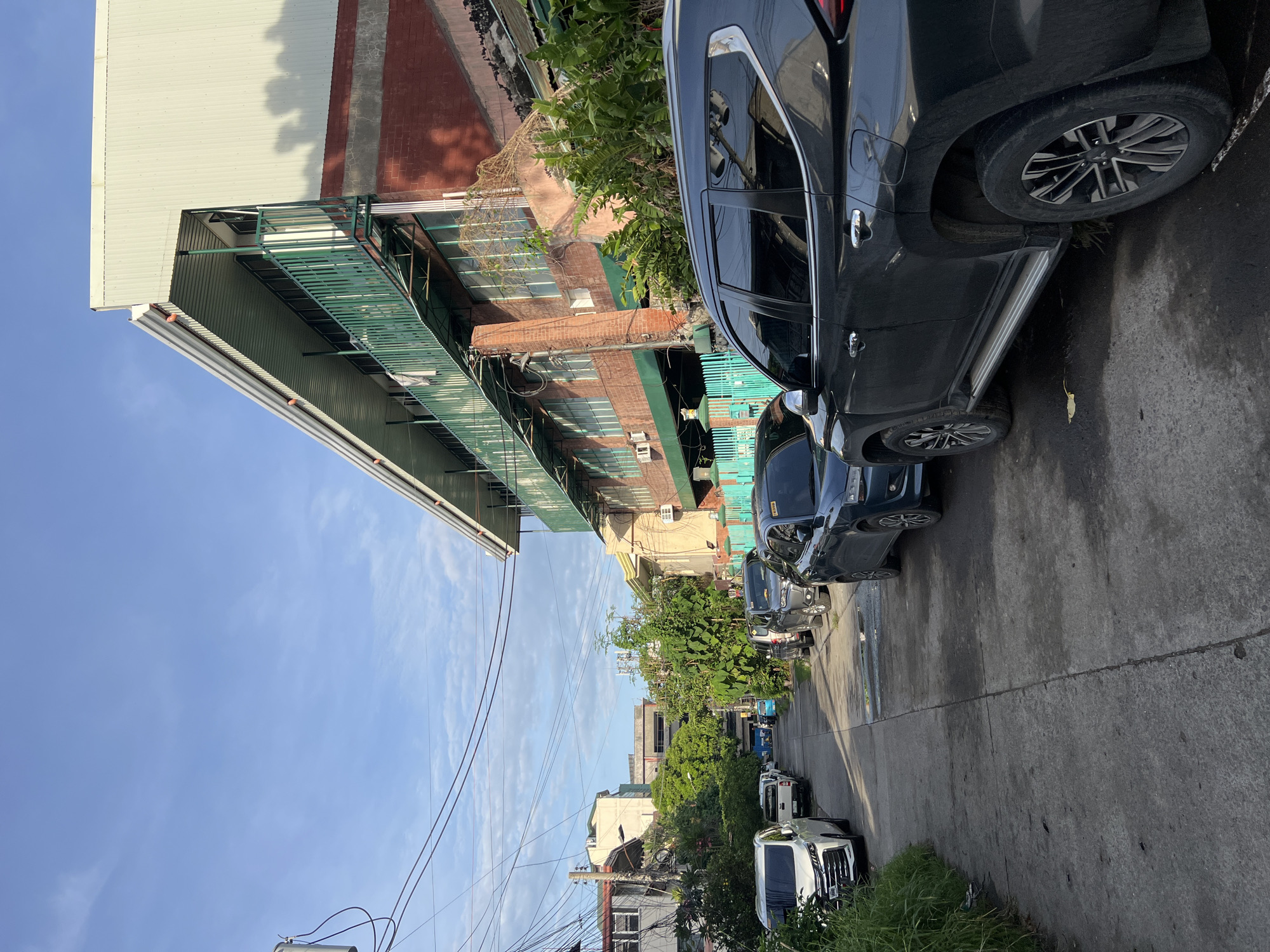 The image depicts a street in Davao City, Philippines. The street is lined with buildings, some of which have a green balcony. Several cars are parked along the side of the road. The sky is visible above the buildings and is blue with some clouds. Power lines are present in the image. Vegetation can be seen in the image, including trees and grass.
