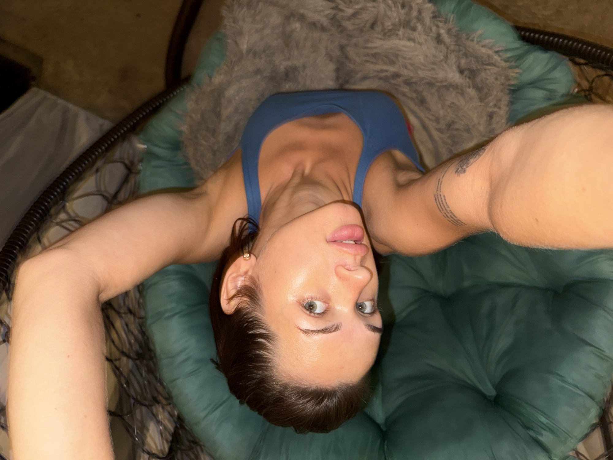 A high-angle shot features a woman lying in a hanging chair with a fuzzy gray blanket. She is wearing a blue tank top. The chair is dark green and appears to be made of plush material. The background is blurry and dark, with some indistinct shapes suggesting an indoor environment.
