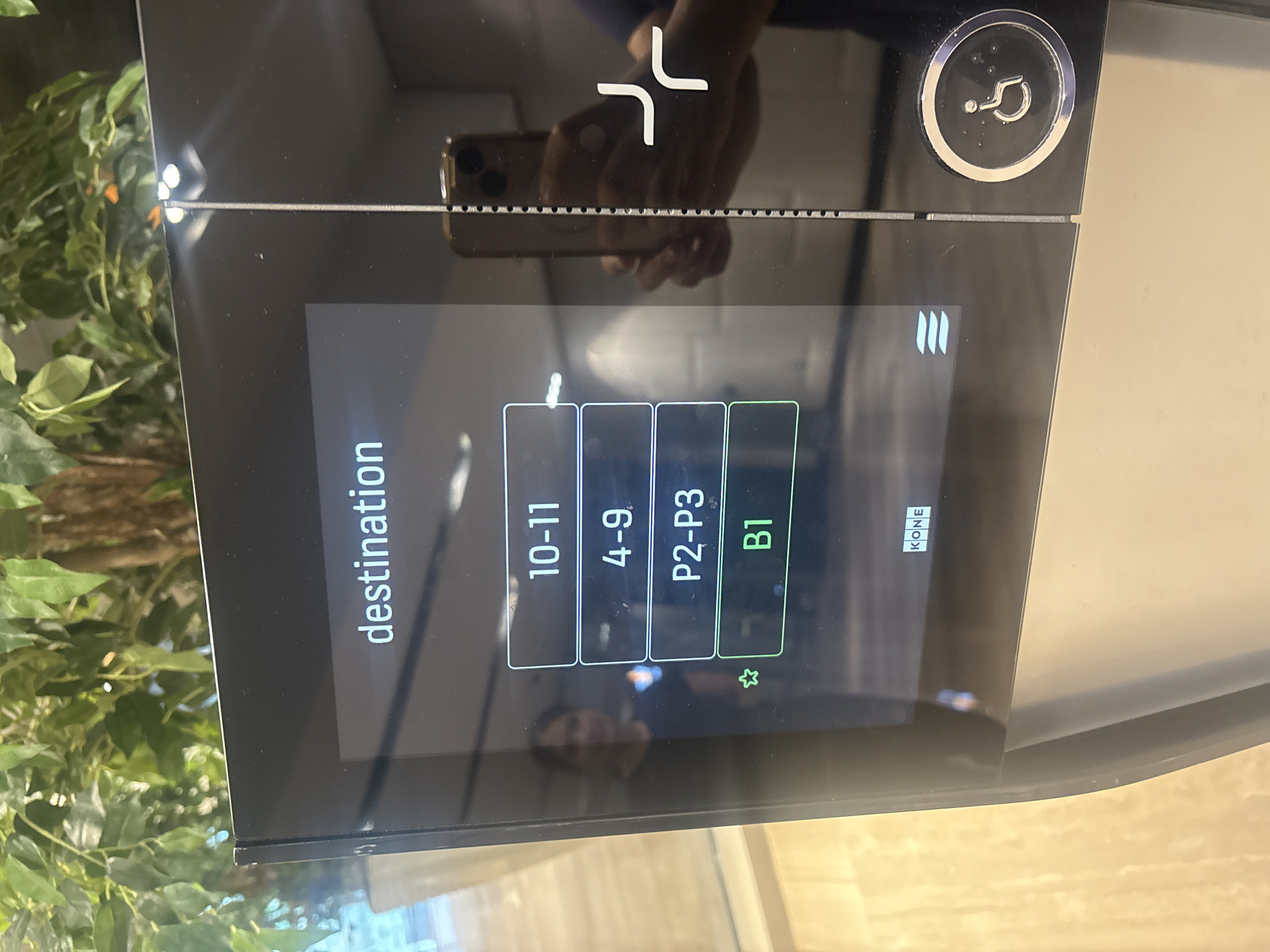 An elevator destination selection panel is shown in the image. The panel displays the word "destination" at the top, followed by a list of selectable floor options: "10-11", "4-9", "P2-P3", and "B1". The "B1" option is highlighted in green, indicating it is the selected destination. A small star icon is visible next to the "B1" option. Below the floor options, the brand name "KONE" is displayed. To the right of the panel, an accessibility symbol (wheelchair icon) is visible within a circular button. The panel is mounted on a dark, reflective surface, which shows reflections of a person holding a smartphone and a portion of a plant with green leaves. The background appears to be an indoor setting with light-colored walls and flooring, possibly a modern building lobby. No specific location in Dubai, United Arab Emirates, can be determined from the image alone.