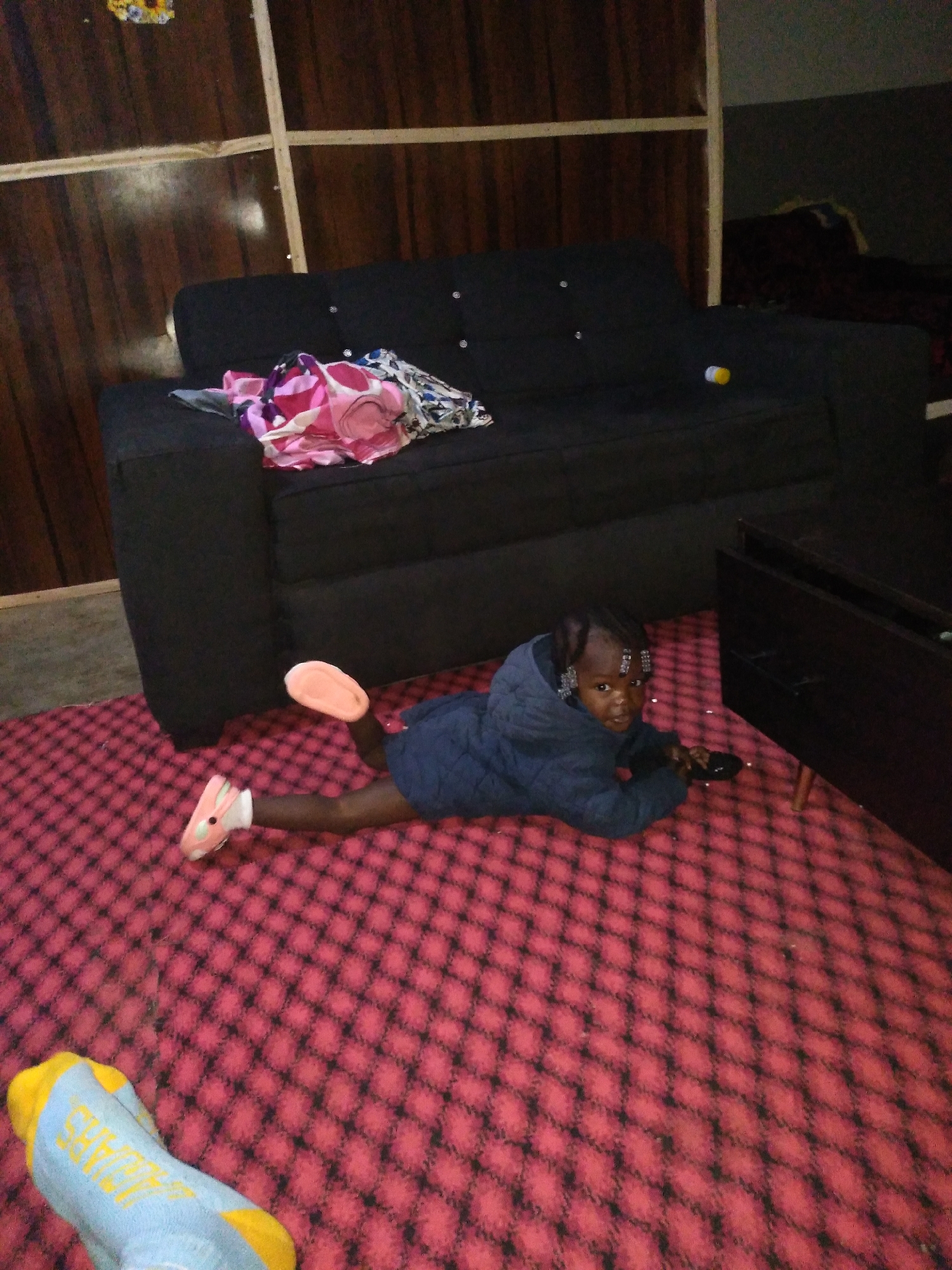 A young Black child is lying on their stomach on a pink and black checkered carpet, facing the camera. The child is wearing a dark blue quilted jacket and pink and white sneakers. Their legs are bent at the knees and their feet are pointed outwards. The child's hair is styled in braids and adorned with sparkly clips. They are holding a dark-colored object, possibly a remote control, in their right hand. To the left of the child, a blue and yellow sock is visible on the carpet. In the background, a black couch is visible, with a pile of colorful clothes on it. Behind the couch, a wooden wall with horizontal and vertical planks is present. To the right of the couch, a dark brown furniture piece with drawers is visible. The scene appears to be indoors, likely a living room.