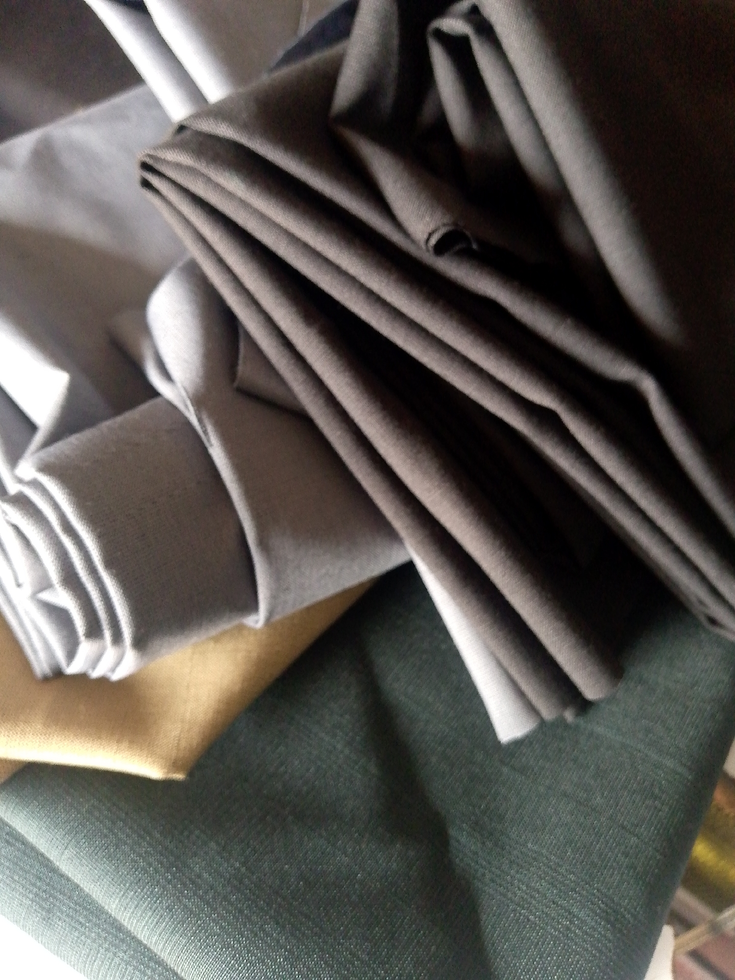 The image shows a pile of folded fabrics in various shades of gray, beige, and dark green. The fabrics appear to be stacked and draped, with visible folds and textures. There are no people or discernible elements of a city environment. The scene is entirely focused on the textiles. There is no information to indicate this scene is located in Jauro Yinu, Nigeria.