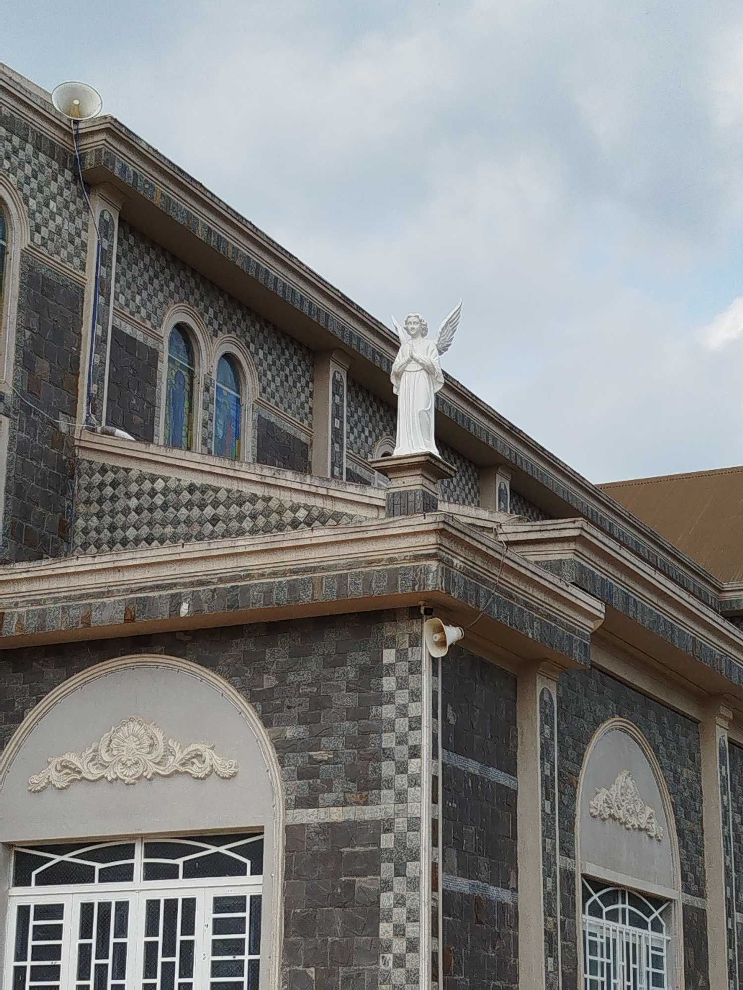 An exterior, upward-looking view captures a distinctive building, likely a church, under an overcast sky. A prominent white statue of an angel, with large wings and hands clasped in prayer, stands on a pedestal at a higher tier of the building's roofline.
The building's facade is constructed from dark grey, rough-hewn stone or tiles, interspersed with sections of lighter, intricate checkered and diamond-patterned tiles. It features multiple levels and rooflines, adorned with light-colored cornices and trim. Arched windows, some appearing to have stained glass in shades of blue and green, are visible on the upper levels. On the lower level, large arched window frames are topped with ornate, shell-like or floral decorative reliefs, and feature white grilles.
Two white, conical public address speakers are mounted on the exterior walls at different heights. A small religious icon or statue is barely discernible within a niche above a lower roof section. The sky is a soft grey with visible clouds, indicating an overcast daytime setting. The architectural style suggests a religious structure, consistent with its likely location in Giãn Dân, Vietnam.