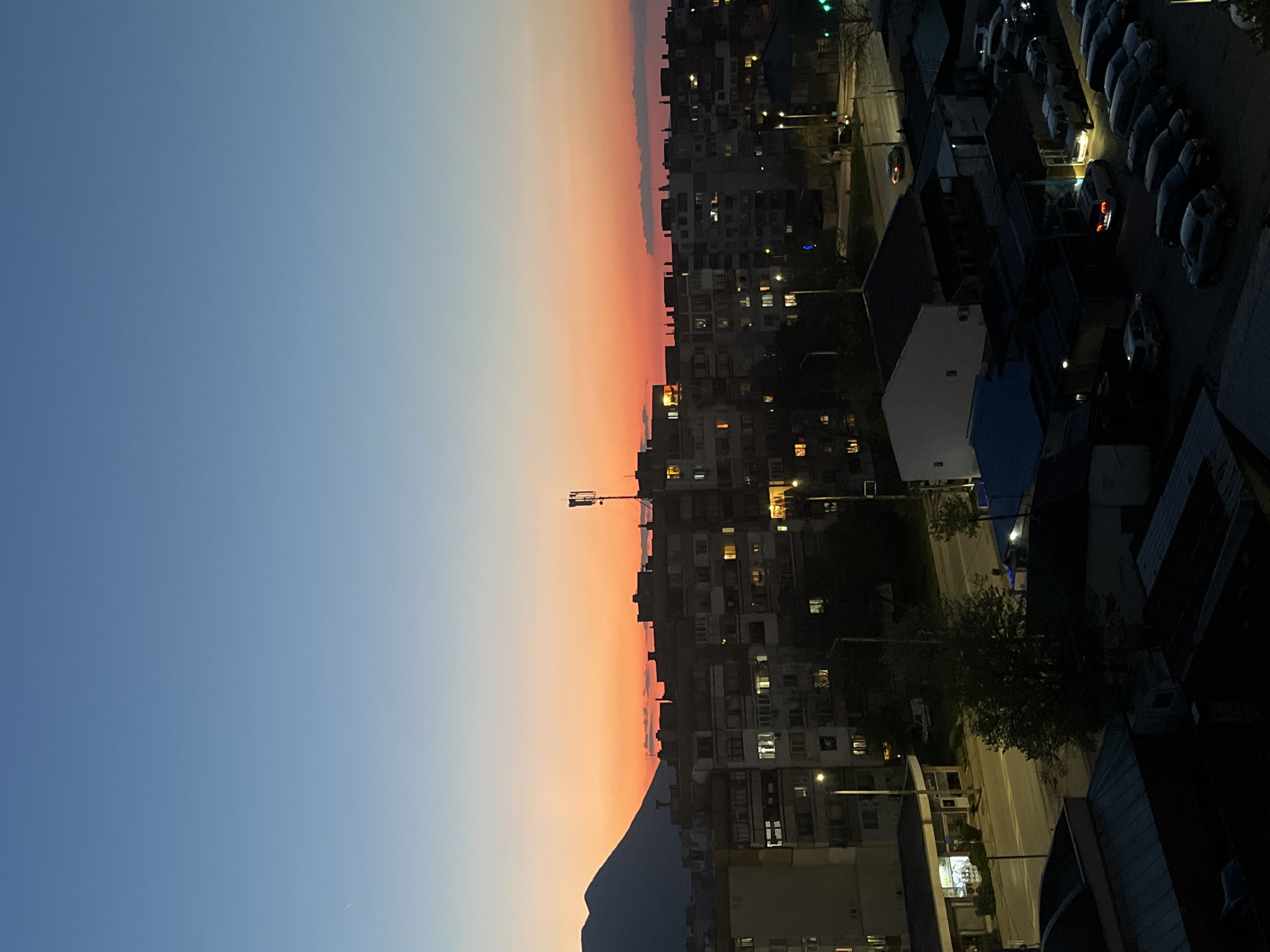 The image shows an evening cityscape in Vratsa, Bulgaria. The sky transitions from blue to orange at the horizon. In the foreground, residential buildings with lit windows stretch along the right side of the frame. A mountain ridge silhouette is visible at the horizon line. Streetlights illuminate the road below, and parked cars are present in a lot.