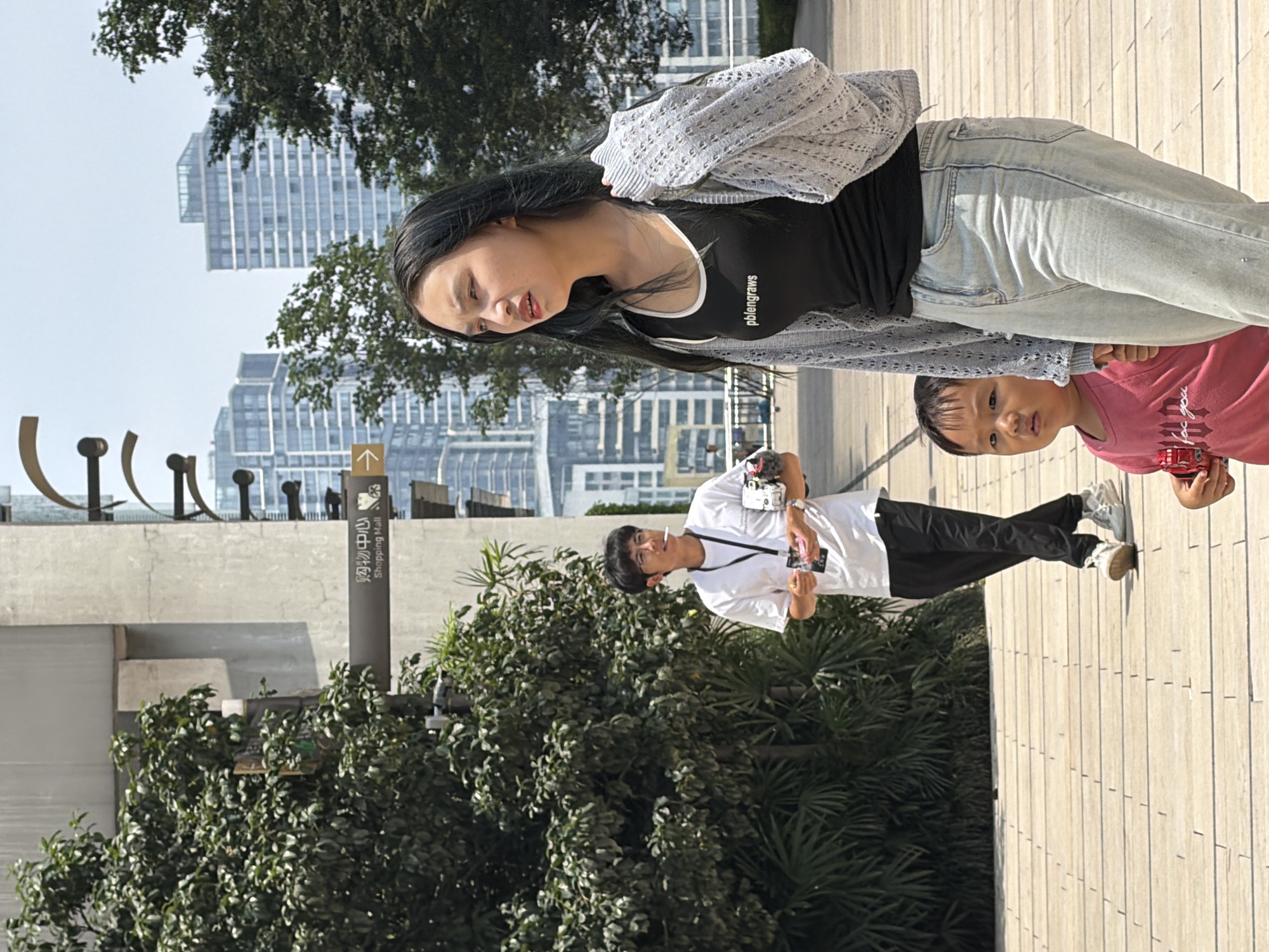 In Yuzhong District, China, a young woman with long dark hair is walking forward while looking to her right. She is wearing a black tank top with white trim that says "pblengraws," light blue jeans, and a light gray knitted cardigan. Beside her and slightly behind, a young child in a pink shirt holds a red toy car and looks toward the viewer. In the background, a man wearing a white t-shirt, black pants, and glasses is walking away from the viewer to the left. He is holding a camera and a pink object in his hands and has a cigarette in his mouth. The three individuals are on a wooden boardwalk. Behind them, to the left, is lush green foliage. Further back, a concrete building with a sign that reads "Shopping Mall" with an arrow pointing right is visible. In the far background, modern skyscrapers rise against a bright sky.