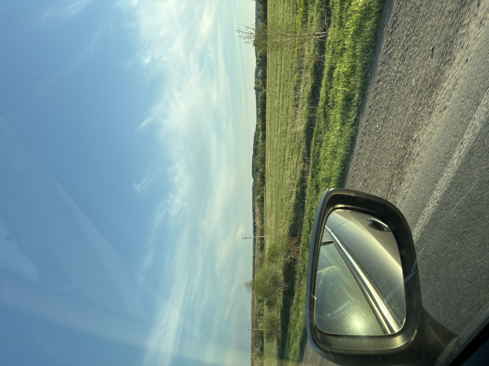 The image depicts a view from inside a vehicle, likely on a road trip. The scene outside the car features a clear blue sky with light clouds. A grassy field extends along the roadside, with trees visible in the distance. A gray side-view mirror is visible, reflecting light and part of the car's interior. The road is asphalt and appears to be in decent condition. The location is in or near the city of Shavkunovo, Russia. There are no people visible.