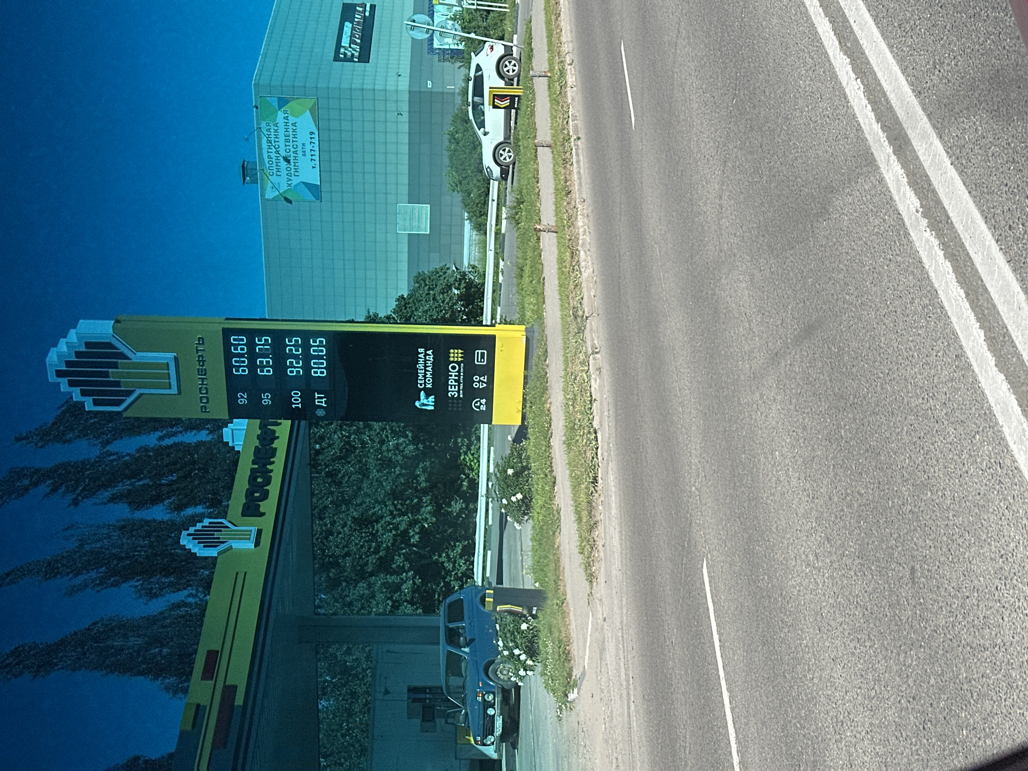 The image shows a gas station in Novoaltaysk, Russia, on a sunny day. A blue car is parked at a gas pump, and a white SUV is driving on the road. A digital sign displays fuel prices. In the background, there is a building with a sign advertising sports and artistic gymnastics. The environment is urban, with trees and a road visible. The photo appears to be taken from a moving vehicle.