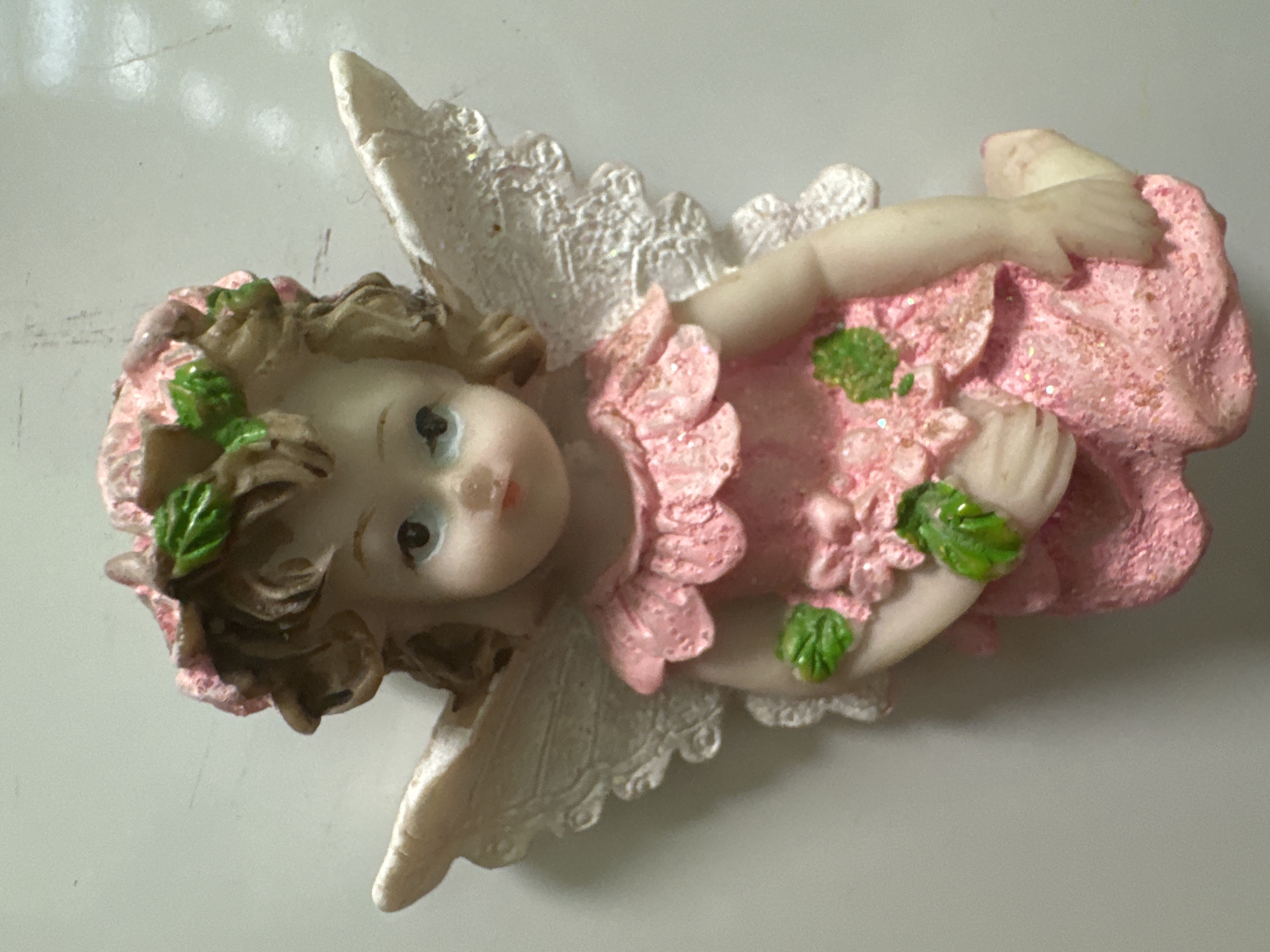 A close-up, overhead shot shows a figurine of a fairy, likely made of resin. The fairy has light skin, light brown curly hair, and is wearing a pink, glitter-covered dress with ruffled trim. Green leaves are adorning the fairy's hair and dress, and there are pink flower embellishments on the dress. The fairy has large, white, lacy wings. The fairy is posed sitting down, with its left leg bent and tucked. The background is a light-colored, textured surface.