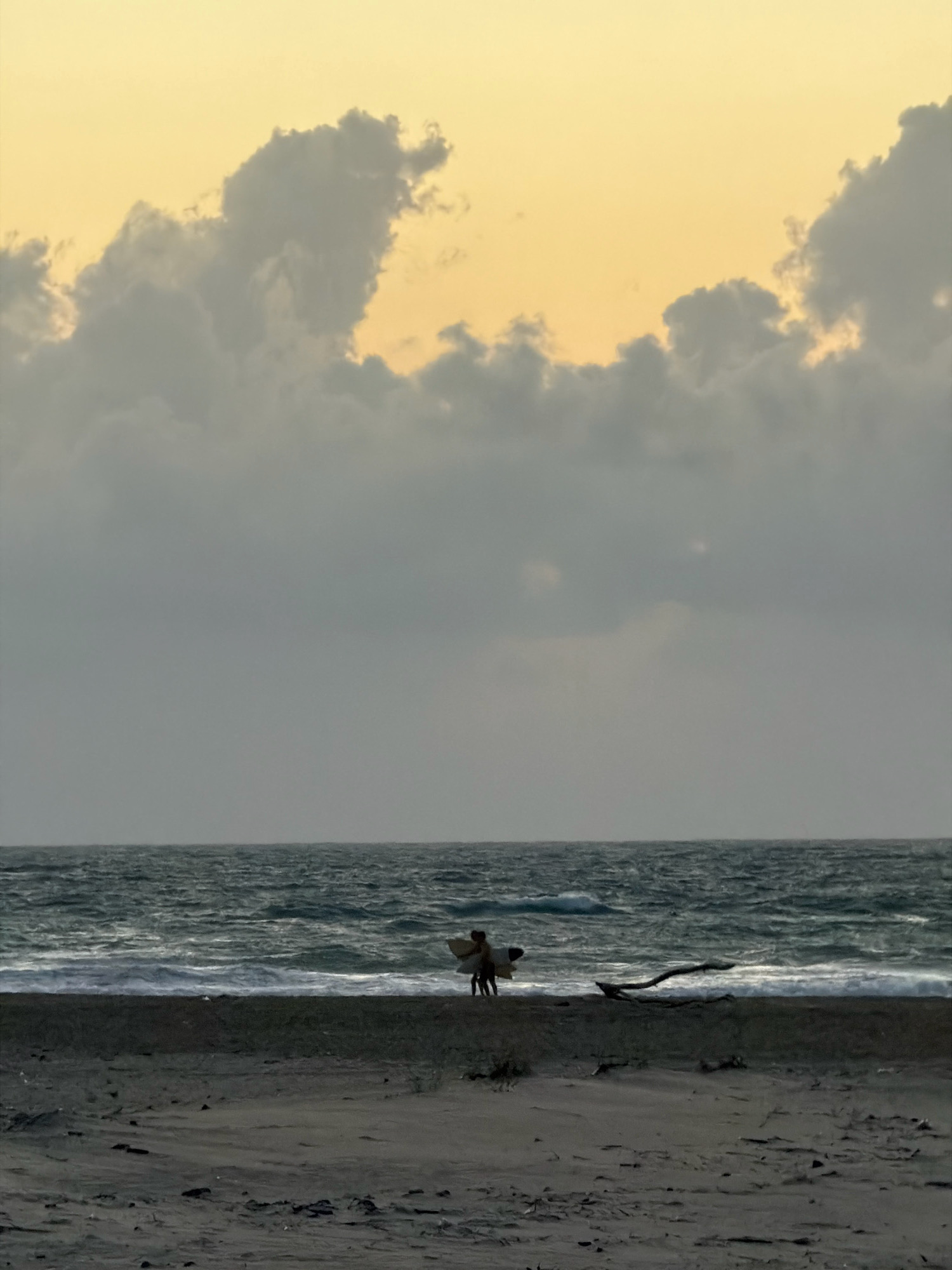 Two silhouetted figures, appearing to be surfers, stand on a sandy beach at the edge of the ocean. One figure holds a surfboard. The sky above is a pale yellow-orange, with heavy, gray clouds obscuring the upper portion of the sky. The ocean exhibits choppy waves, and the beach is sandy with some debris scattered across it, including a prominent piece of driftwood. The scene appears to be set during a time of low light, possibly dawn or dusk. This scene is located in Palmachim, Israel.