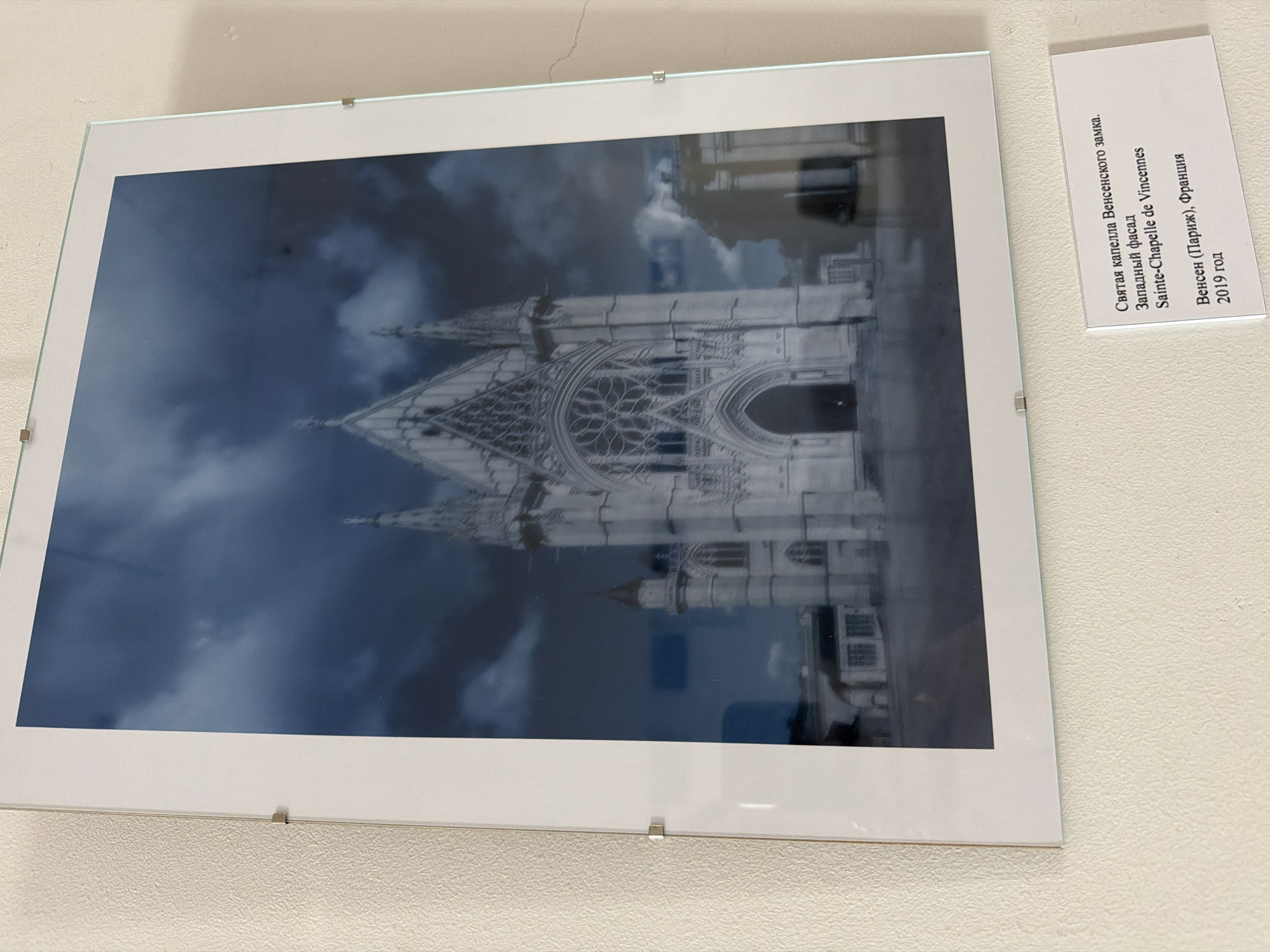 The image is a photograph of a framed artwork hanging on a textured, light-colored wall. The artwork itself is a picture of a gothic-style building, likely a chapel or church, with intricate architectural details, including a large rose window above an arched entrance and spires. The sky in the photograph is dark and cloudy, giving the scene a dramatic mood. Below the artwork, a small white card is attached to the wall, displaying text in Russian and French. The text identifies the building as the Sainte-Chapelle de Vincennes, with the description "Western facade," and the location as "Vincennes (Paris), France," dated 2019. The photograph is taken from a frontal perspective, slightly angled upwards. There is no indication of the location being in Nizhny Novgorod, Russia.