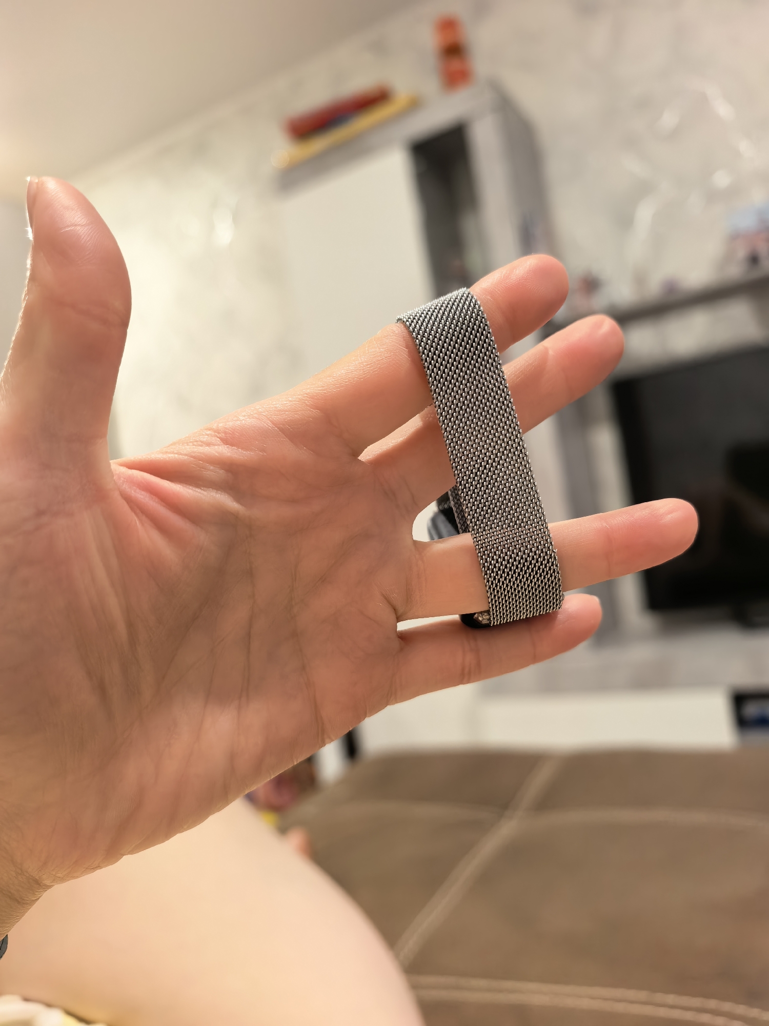 A hand holds a watchband in front of a blurry background. The hand is light-skinned and the watchband is silver and appears to be made of a mesh material. The background shows a white cabinet, a dark television screen, and a brown couch. The location is not discernible in the image, so it is not possible to verify if this image was taken in Sterlitamak, Russia.