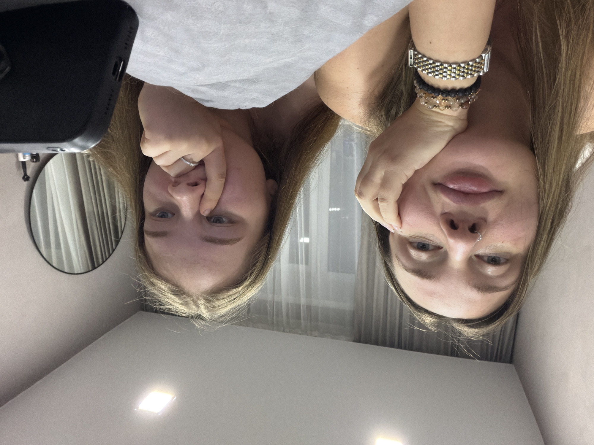 Two young women are pictured in an indoor setting. The woman on the left has blonde hair and is wearing a ring on her finger. Her hand is near her nose. The woman on the right has similar features, with wrist jewelry visible. A black mobile phone is visible in the upper left corner. There is a mirror on the left side of the image. The walls are white. The location is not explicitly stated in the image and would require further information to verify the city of Chaikovsky, Russia.