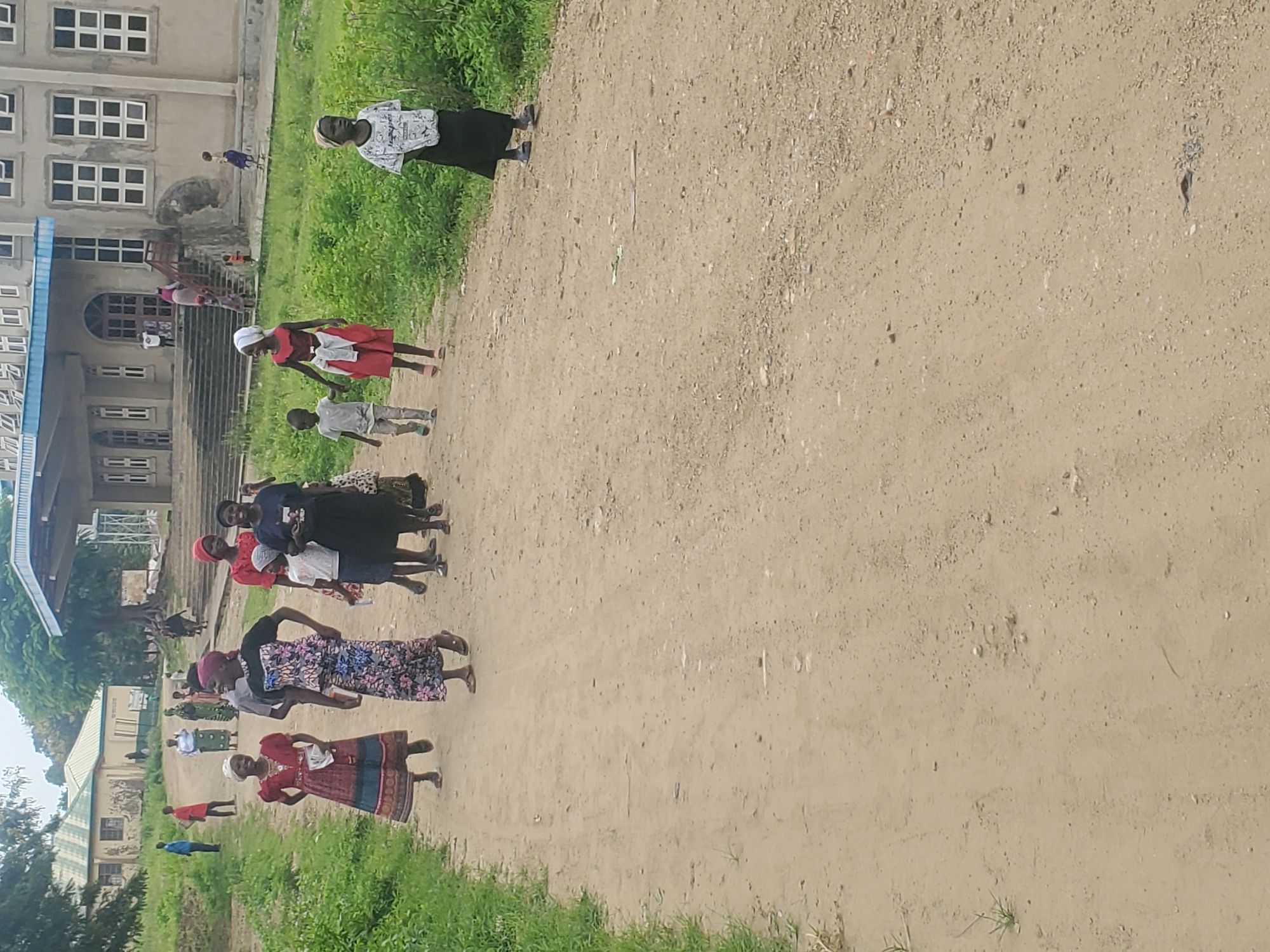 The image shows a group of people, including men, women, and children, walking on a dirt road in front of a large, light-colored building. The building appears to be a school or a church, with multiple windows and a prominent entrance with stairs. The area is surrounded by green vegetation and trees. The people are dressed in various colorful clothing, and some of them are carrying bags or other items. The atmosphere seems to be relaxed and casual. The location is identified as Kabwir, Nigeria, based on the provided context. The scene depicts a community gathering or procession, possibly for a religious or educational event.